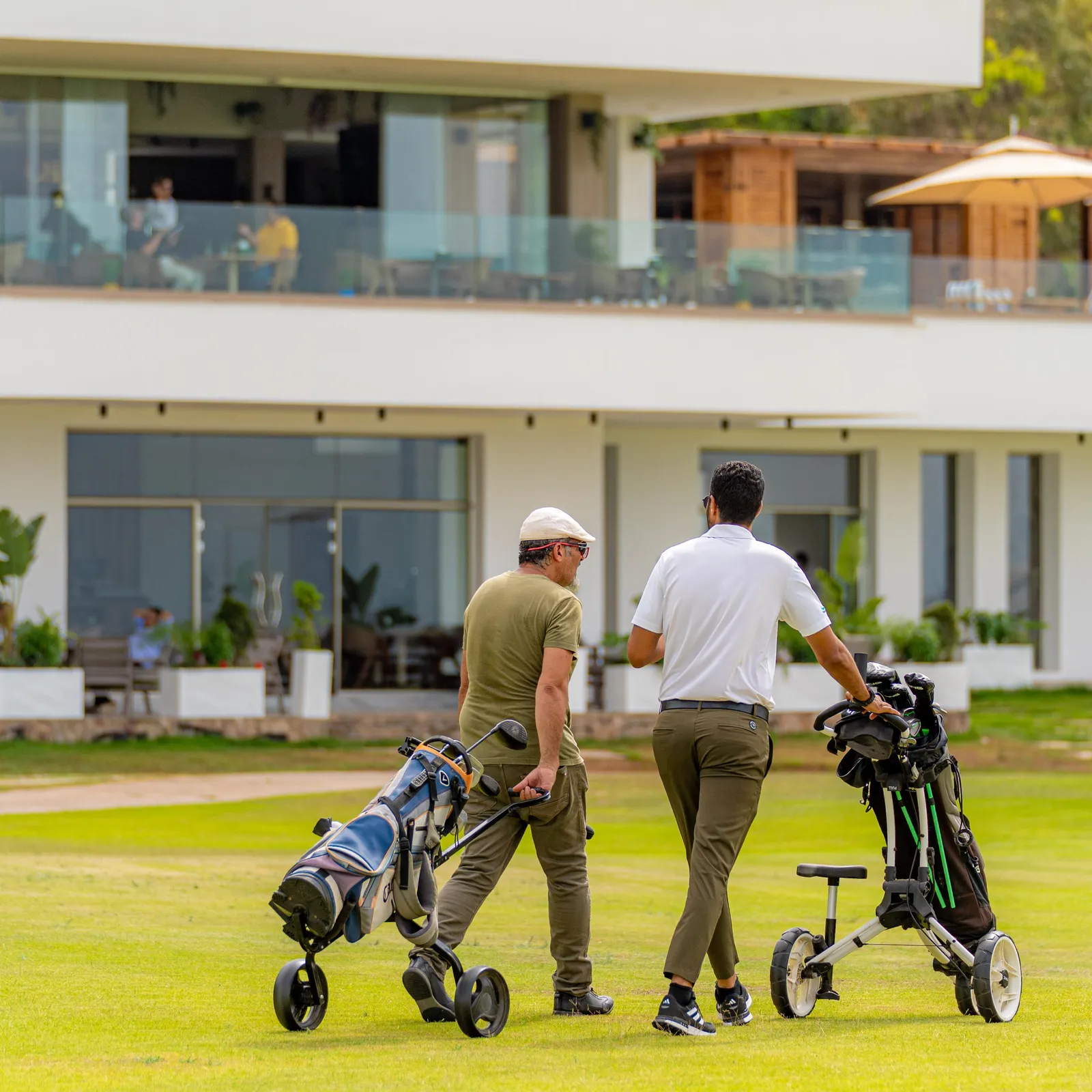 Two men walk on a golf course with club bags, in front of a modern building with large glass windows.
