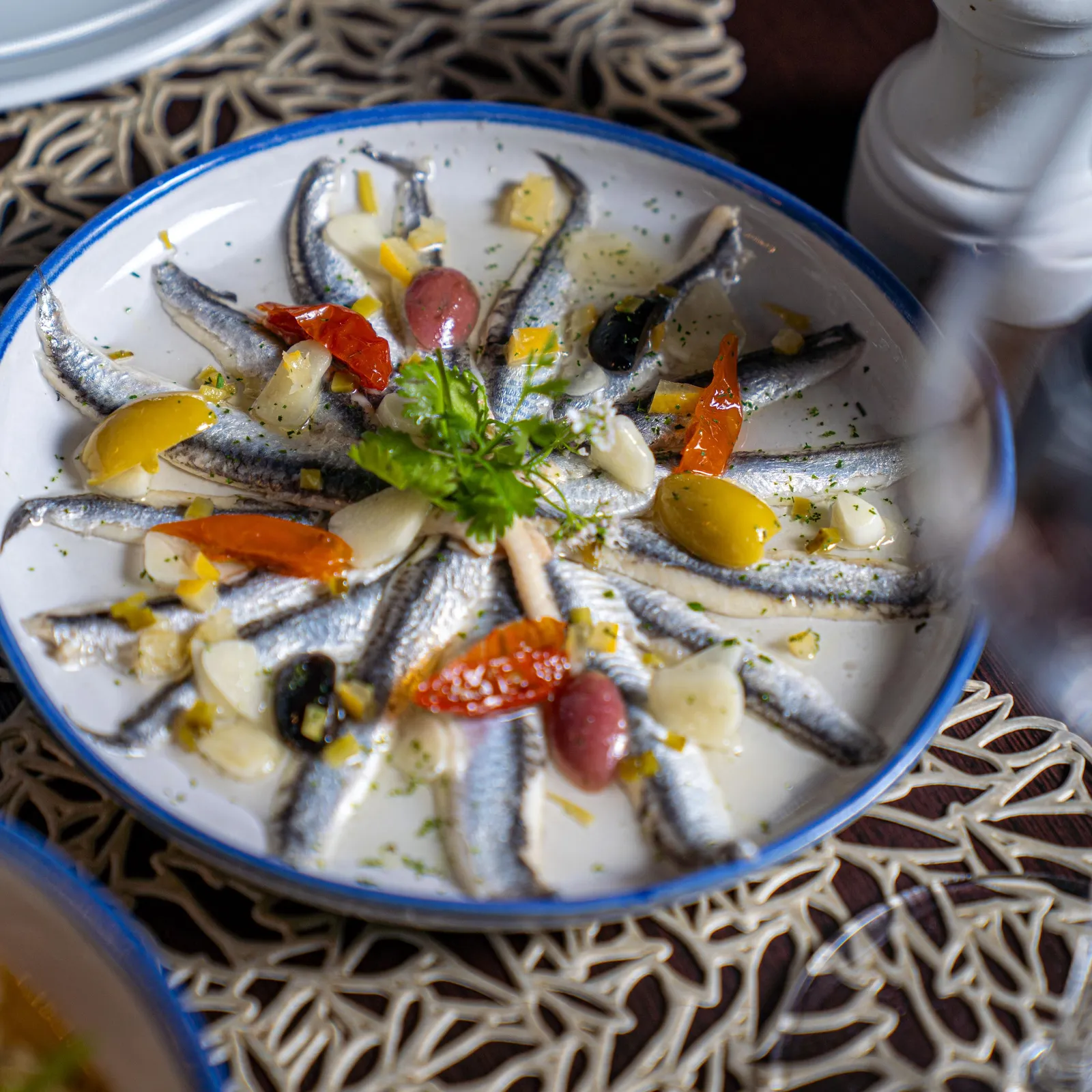 A plate of grilled sardines with vegetables and olives, served on a white plate with a blue rim on a table covered with a patterned tablecloth.