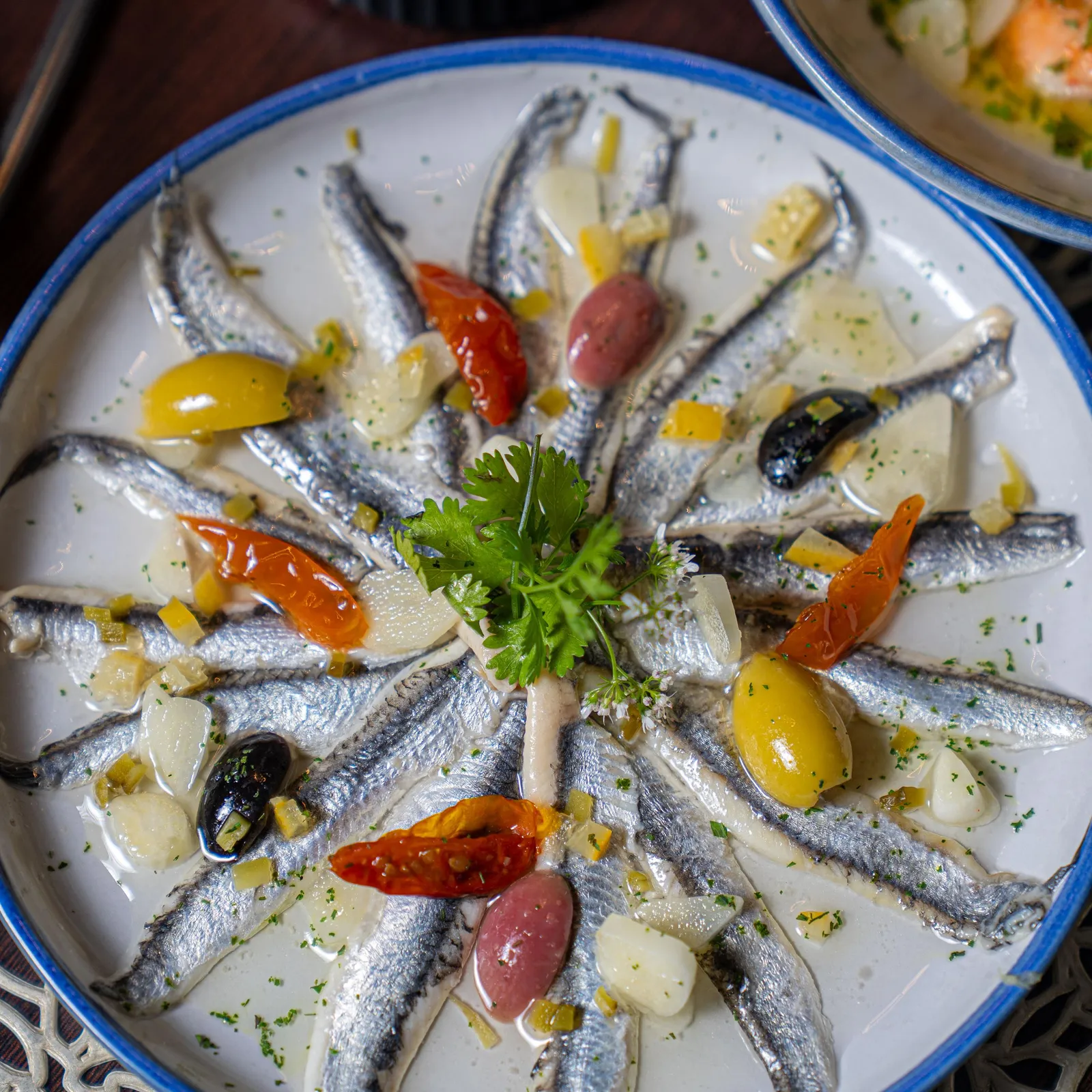 A dish of marinated sardines with olives, sun-dried tomatoes, capers, and herbs, served in a white plate with a blue rim.