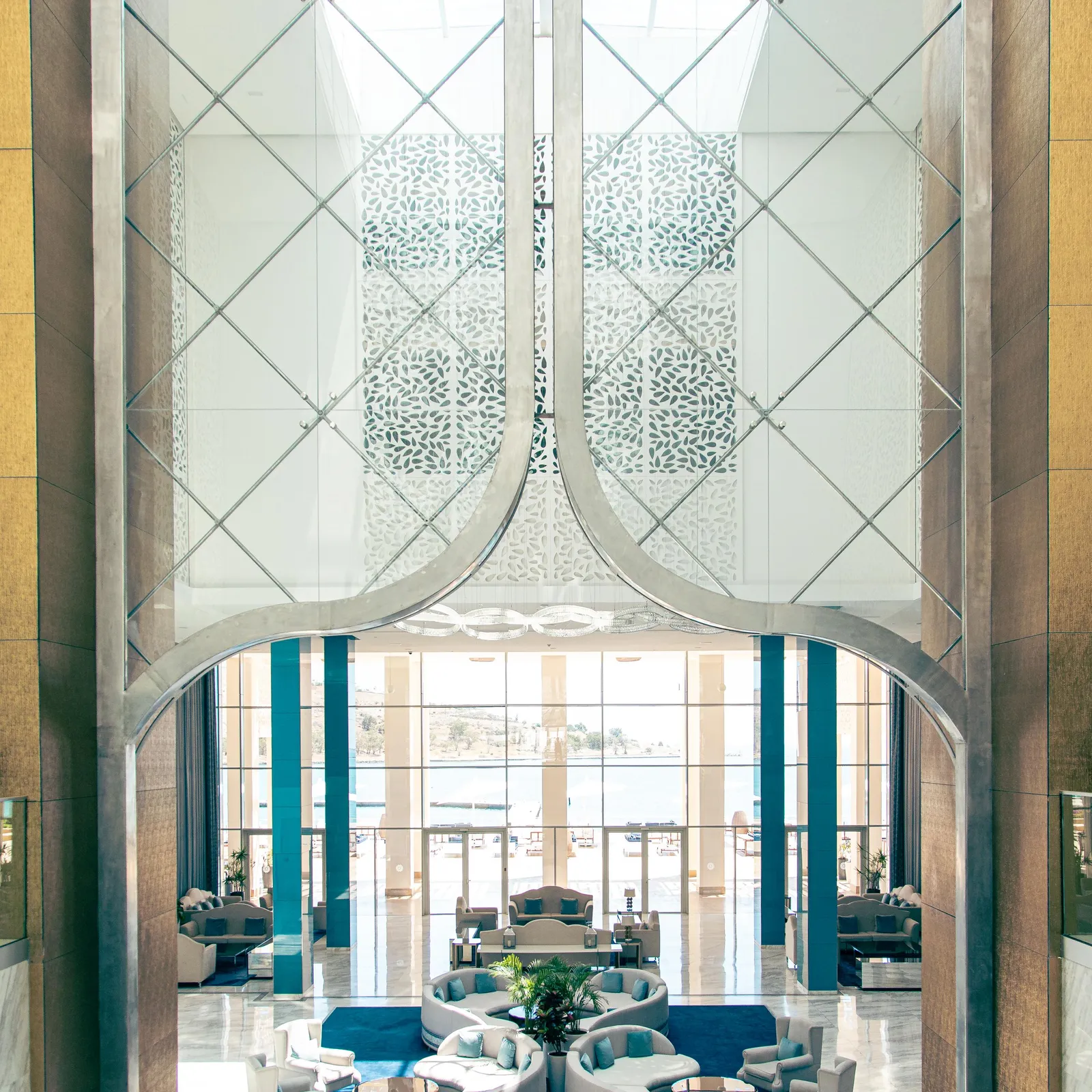 View of an elegant living room through a glass and metal structure with geometric patterns, showing sofas and armchairs arranged around coffee tables