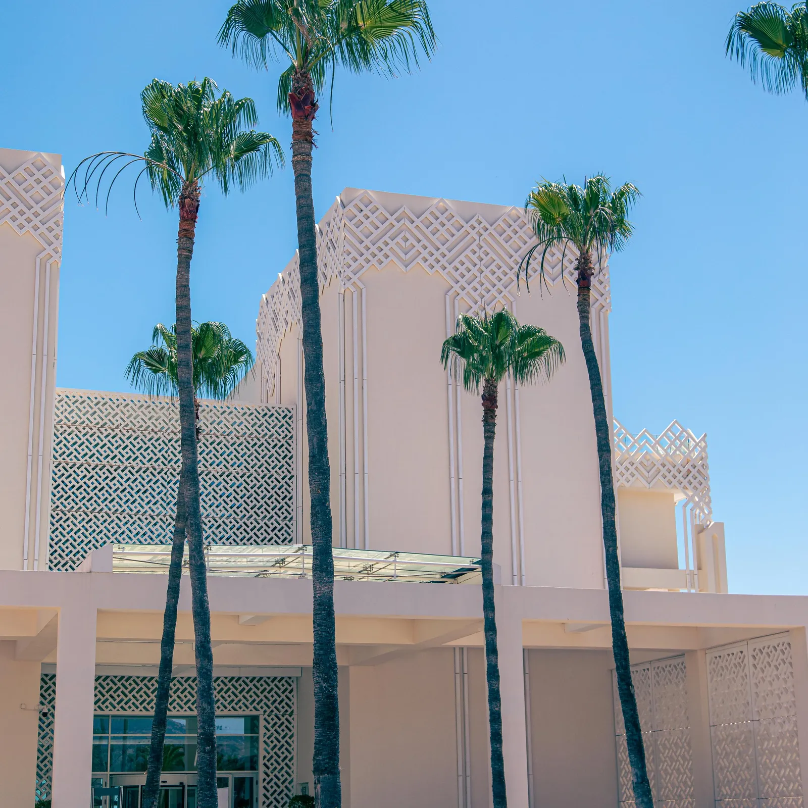 A beige building with geometric patterns and palm trees in front of a fountain.