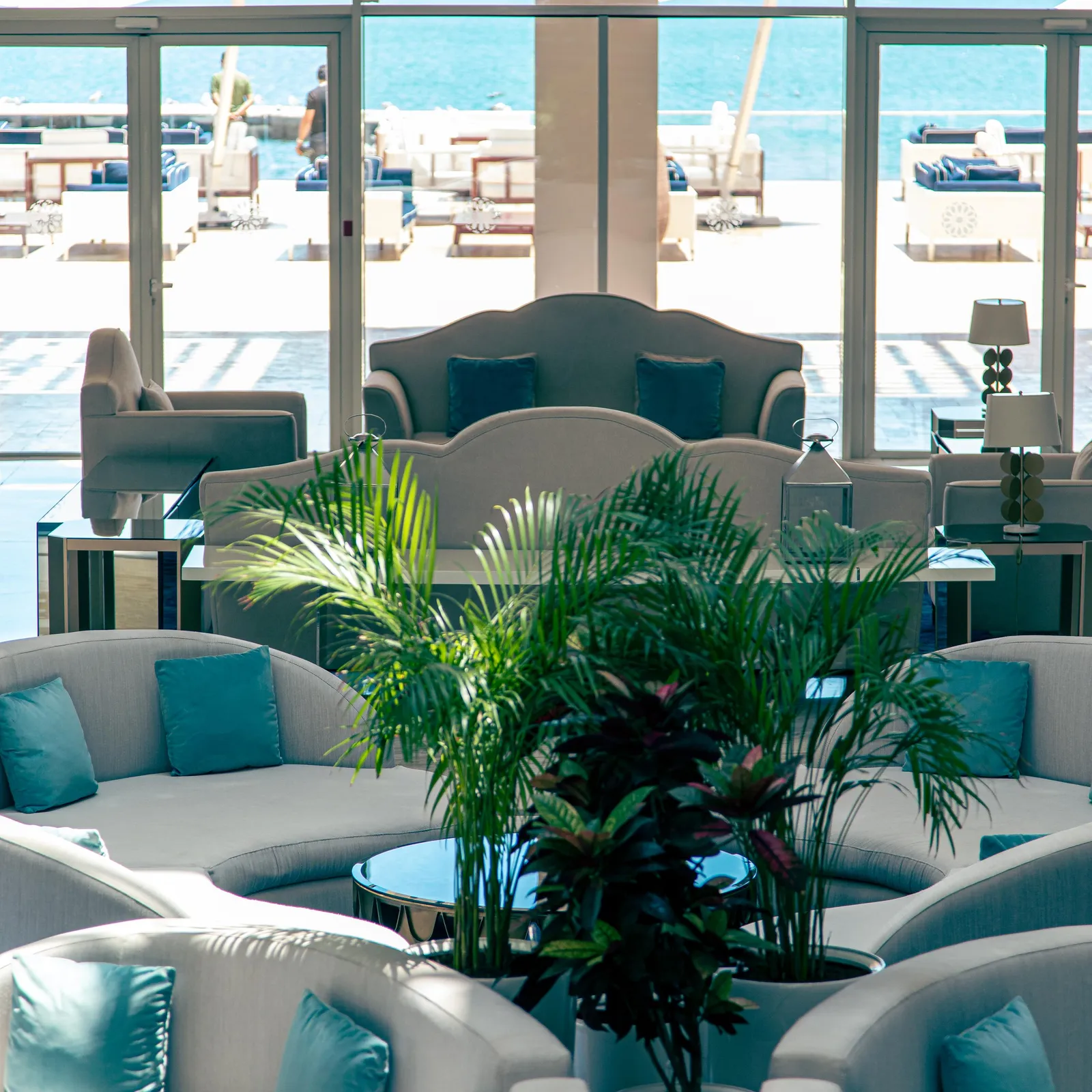 A living room with gray sofas and blue cushions, a green plant in the center, and a view of a pool and the sea through large glass windows.