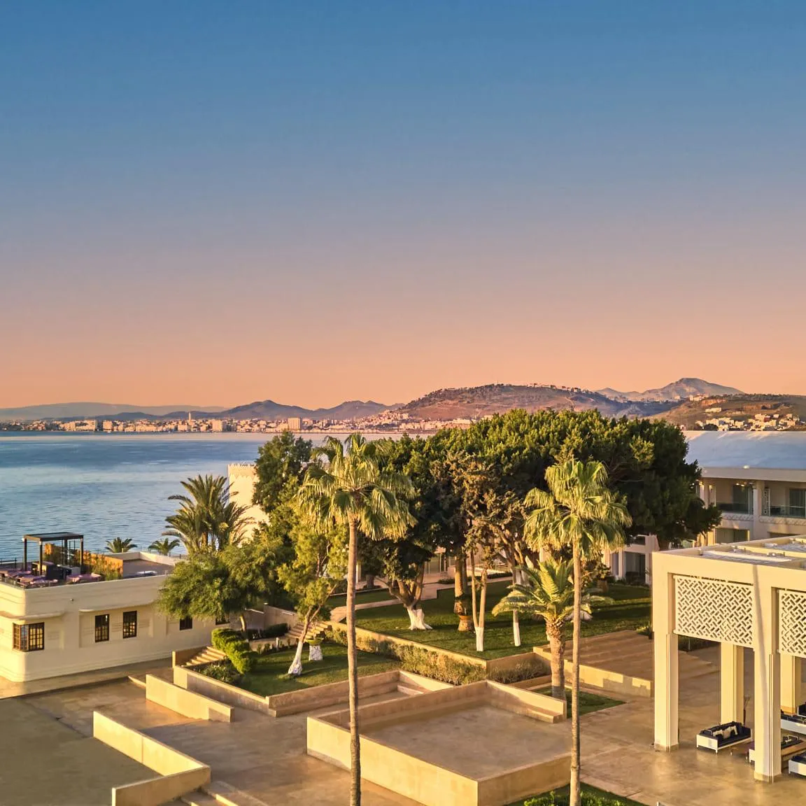 View of a modern seaside architectural complex with white buildings, a tower, palm trees, and a calm sea at sunset