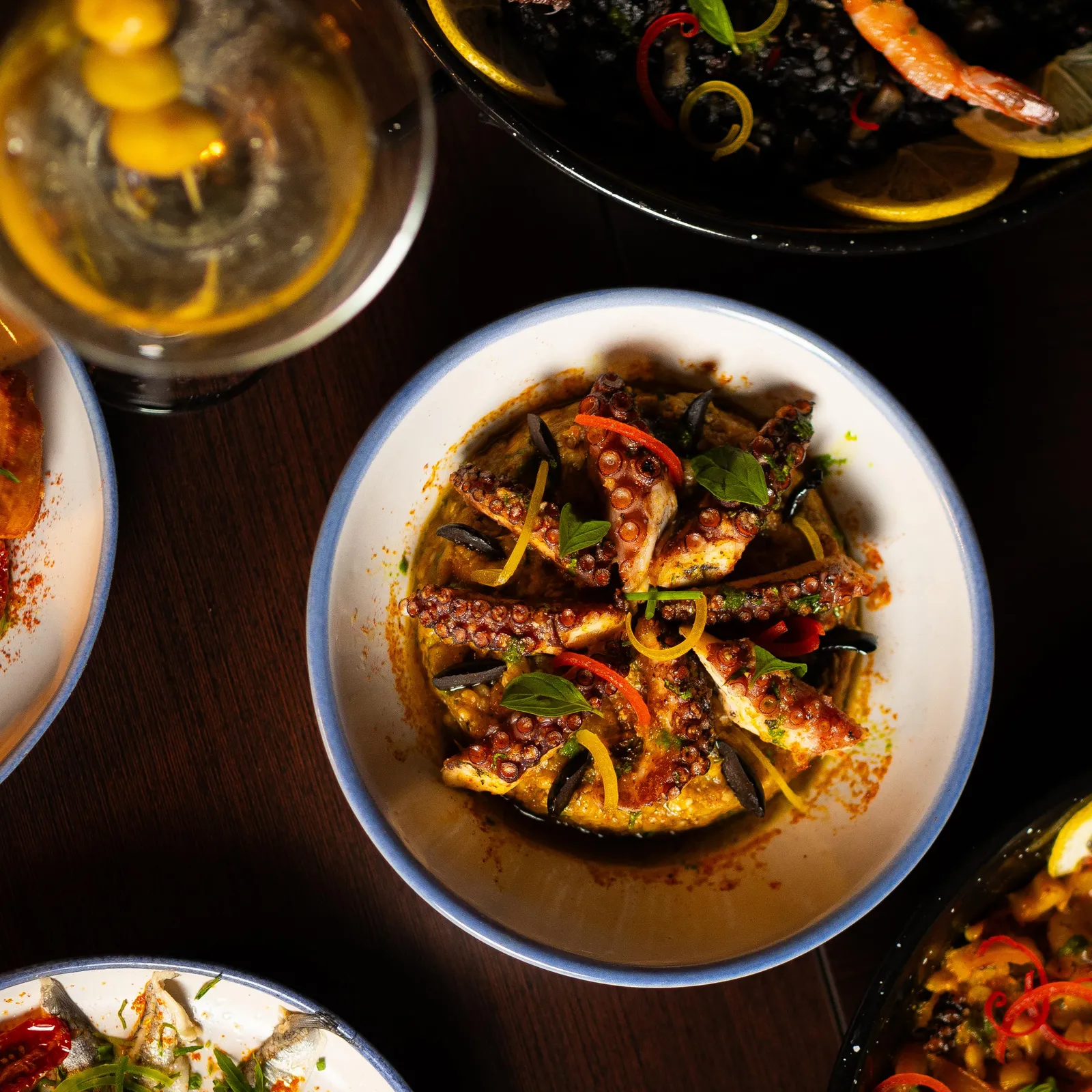 A grilled squid dish with vegetables and herbs, served with other seafood plates and a drink in a glass.