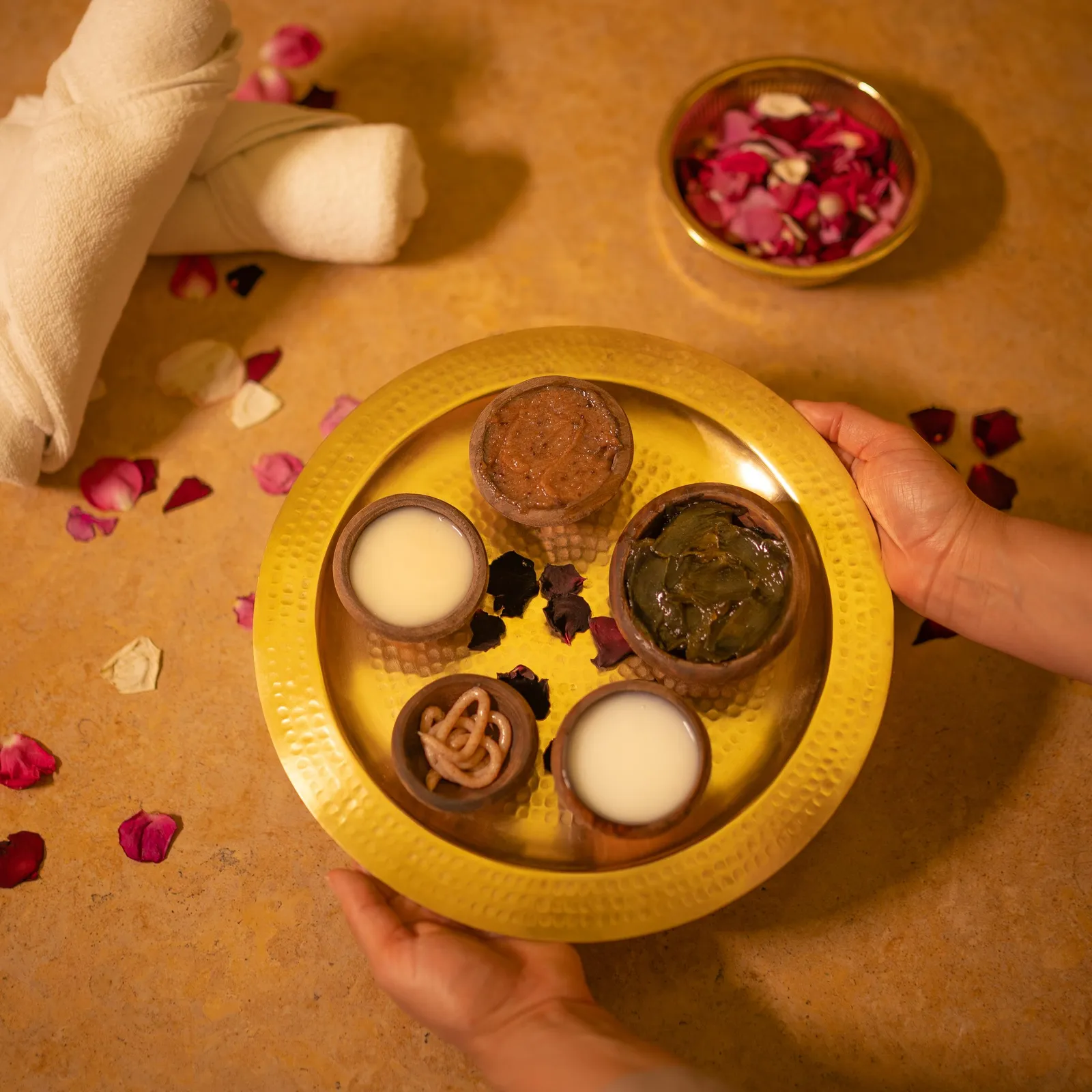 A yellow tray with several small containers holding various substances is held by two hands, with rose petals scattered around.