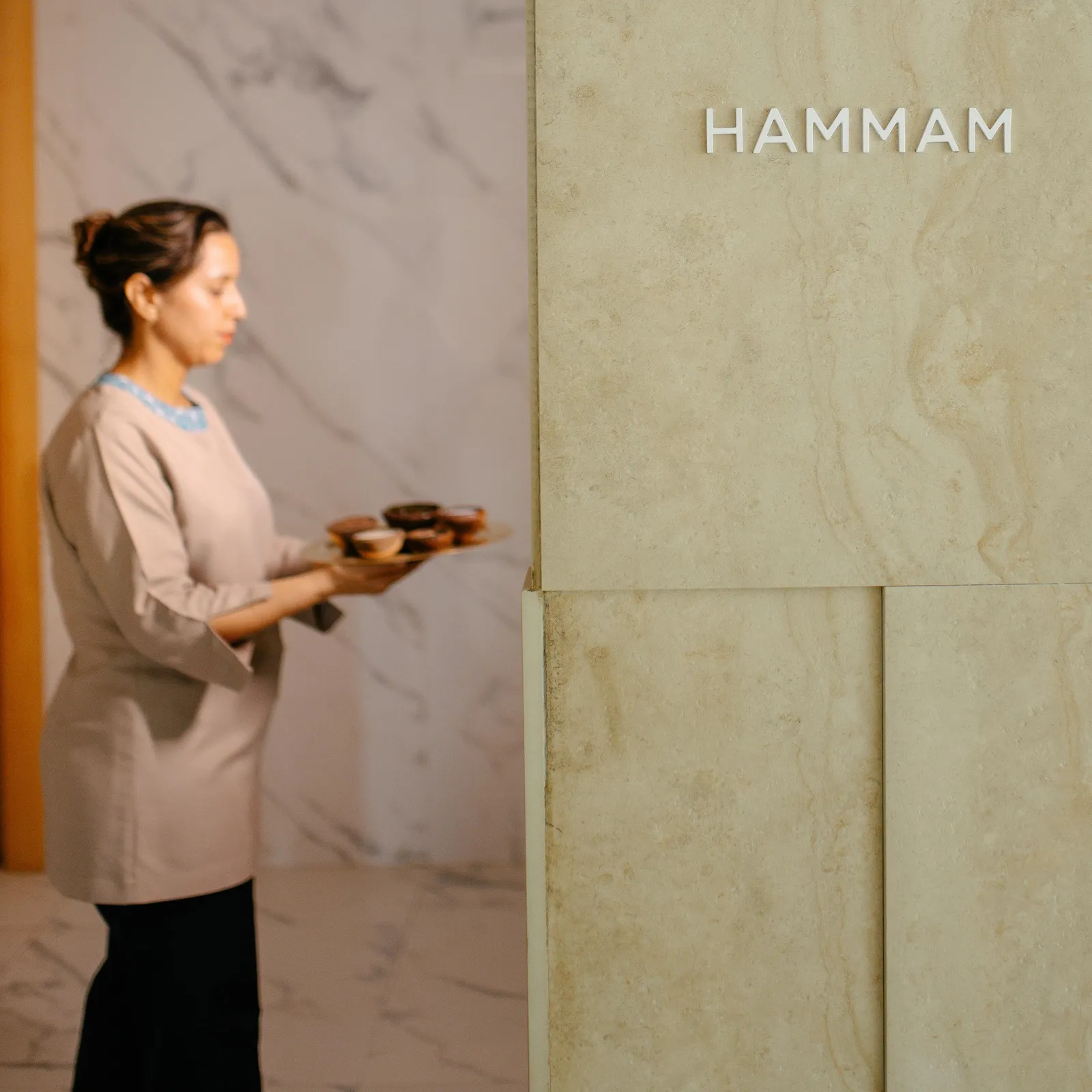 Woman in traditional attire carrying a tray in an interior with stone walls and the inscription "HAMMAM"