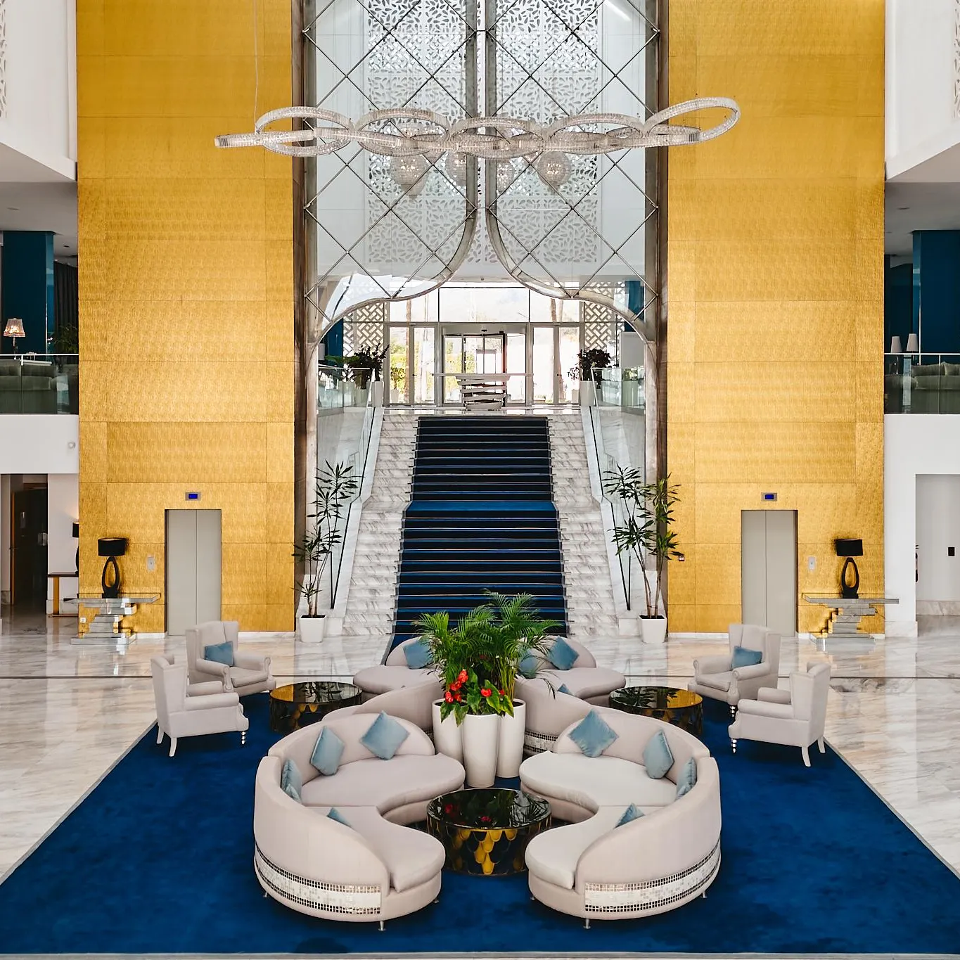 Hotel lobby with white sofas, green plants, a central staircase, and a large suspended chandelier.