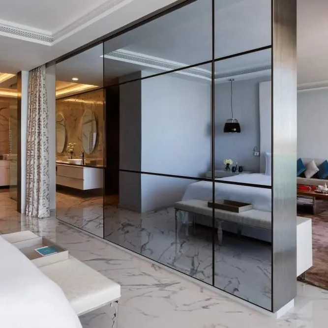 Modern interior with a mirror separating a bedroom and a living room, featuring white furniture and a seating area with a sofa and armchair.