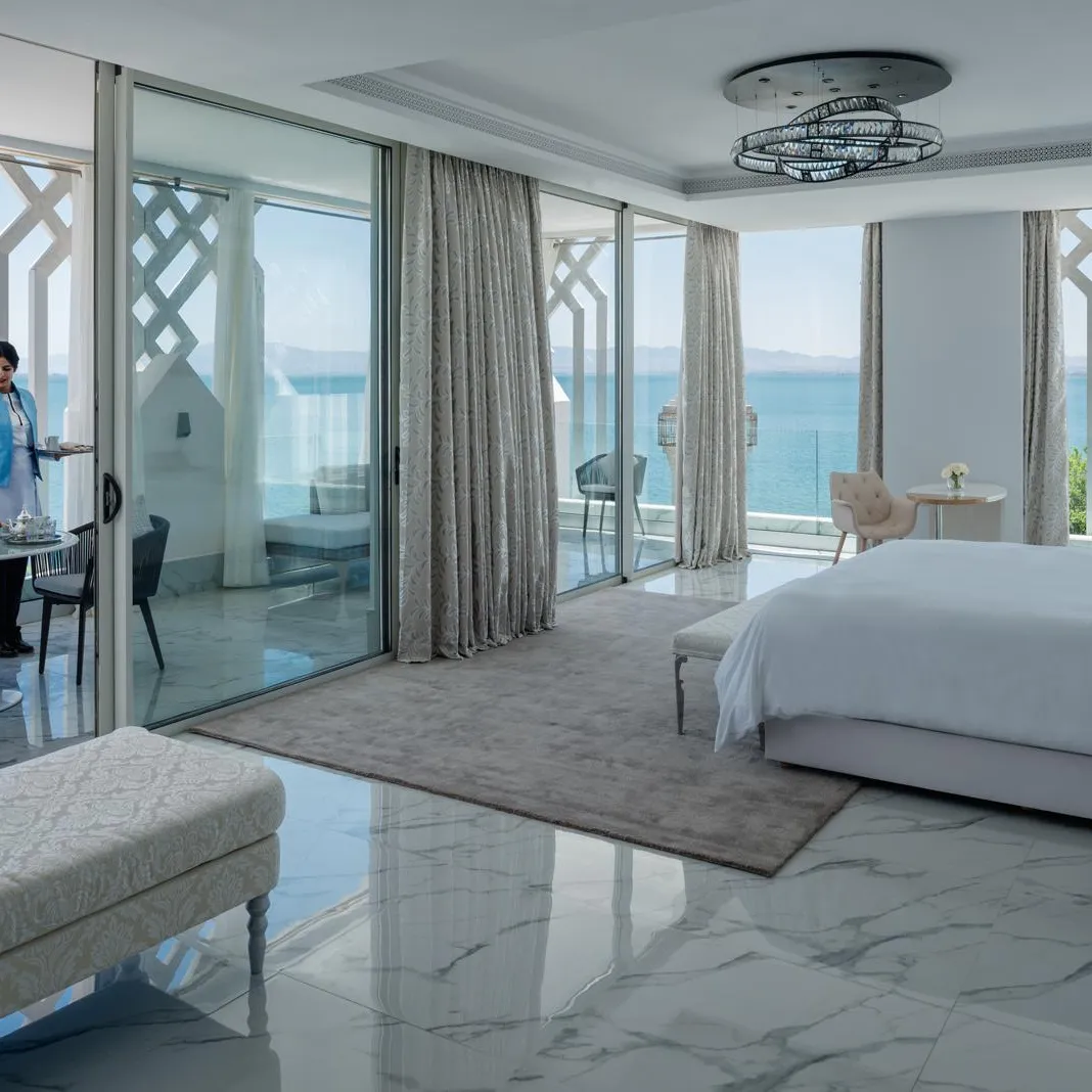 Luxurious hotel room with sea view, a bed, a sofa, a dining table and a person serving a tray