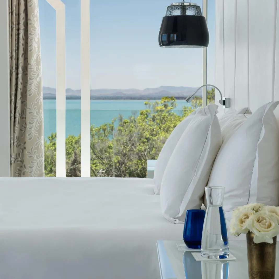 View of a room with a white bed and pillows, a bedside table with vases and flowers, and a large window overlooking a blue sea with mountains in the distance.