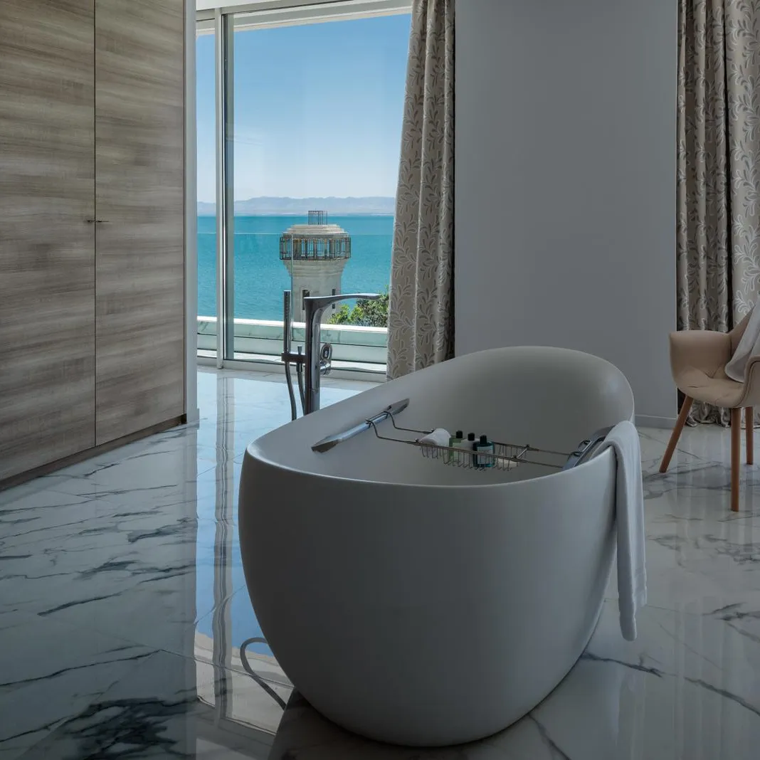 White bathtub with towels placed on it, located in a room with a sea view and an armchair covered with a towel.