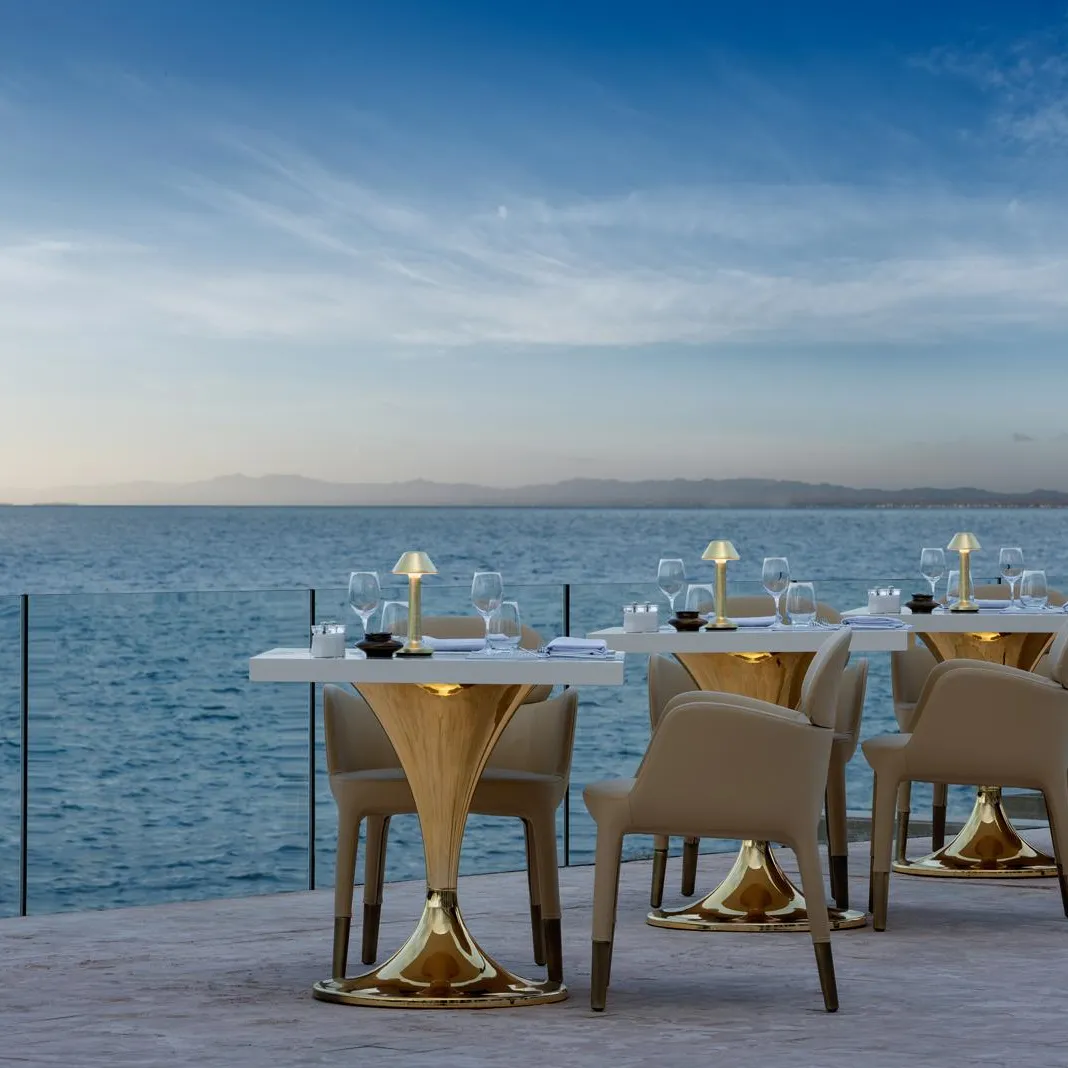 Tables and chairs arranged on a terrace facing the sea with glasses and napkins, under a clear sky.