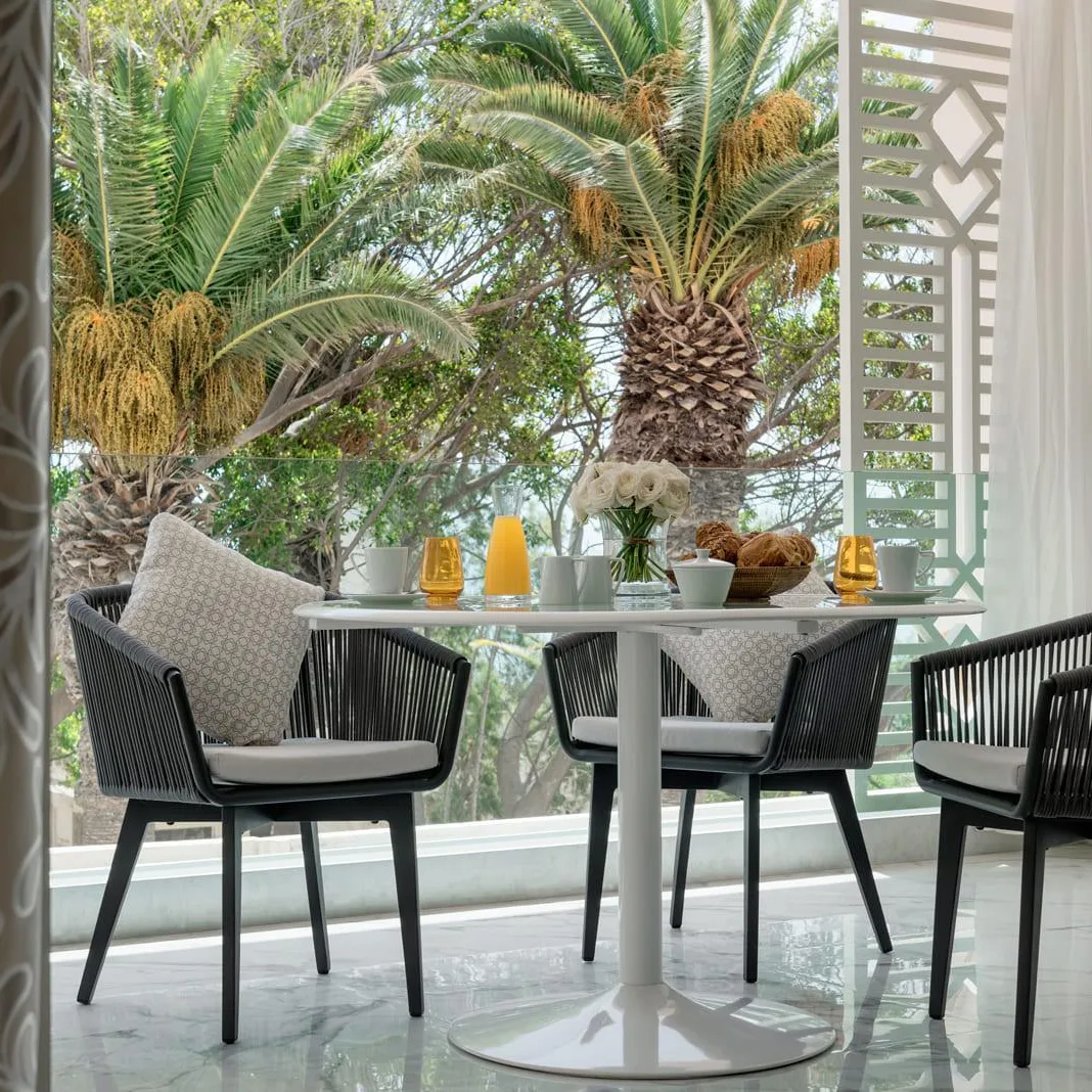 Round table with four black rattan chairs and gray cushions, seen from an open door, with plants and decorative elements on the table in the background.