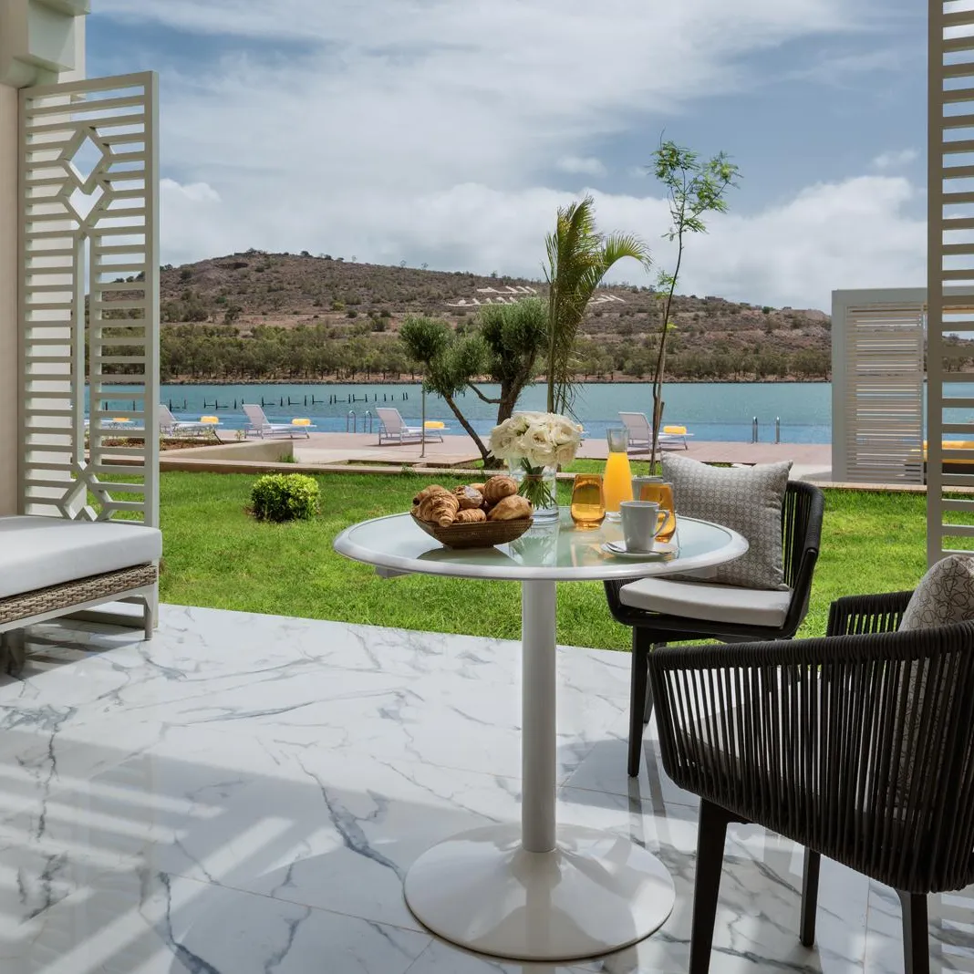 Terrace with table, chairs, and sofa overlooking a lawn and a lake with sun loungers and mountains in the background.