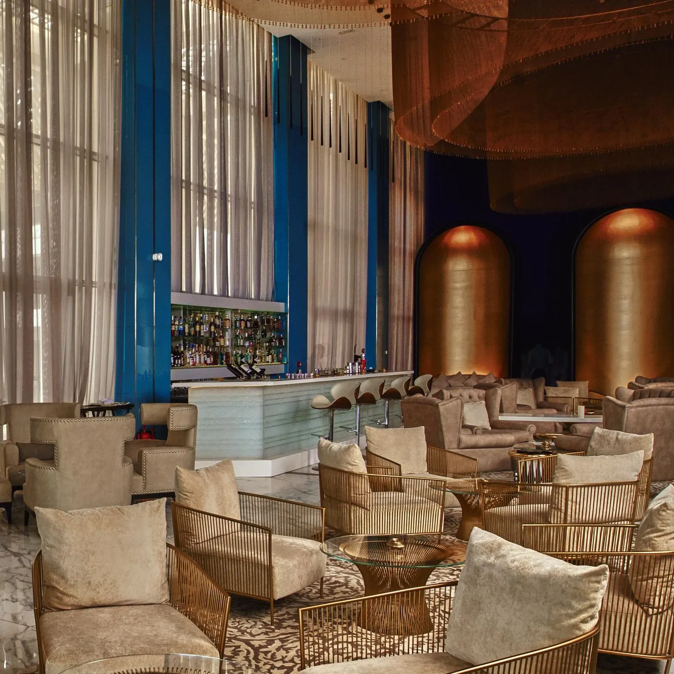 An elegant lounge with beige velvet armchairs, glass and metal coffee tables, a well-lit bar, and sheer curtains in front of large windows.