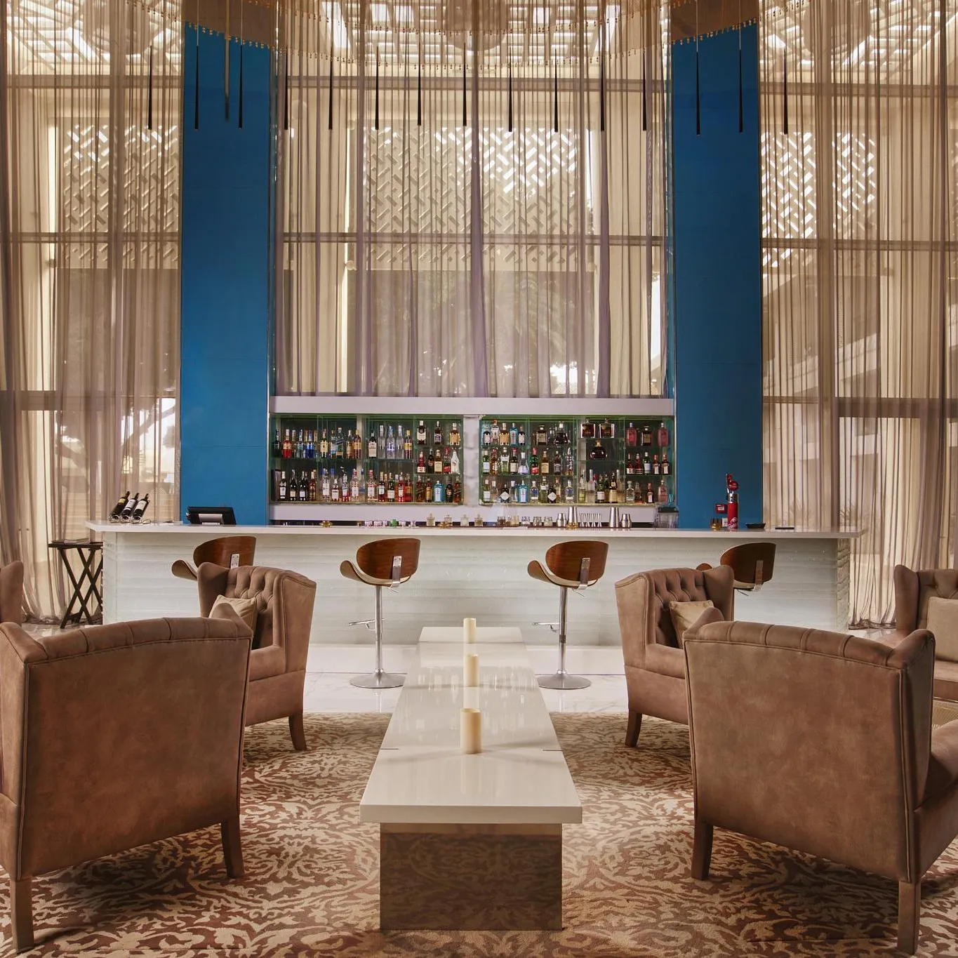 A living room with leather armchairs around a coffee table, facing a bar with shelves filled with bottles, behind large windows with sheer curtains.