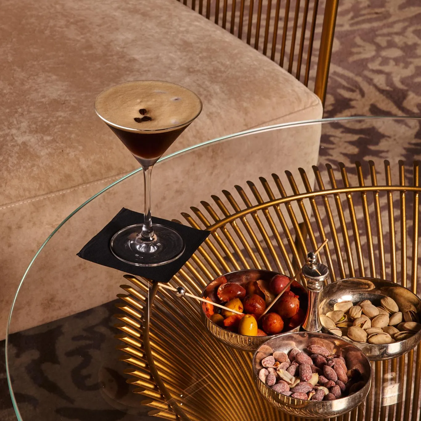 A glass of cocktail placed on a low glass and gold metal table with bowls of dried fruit and pistachios.