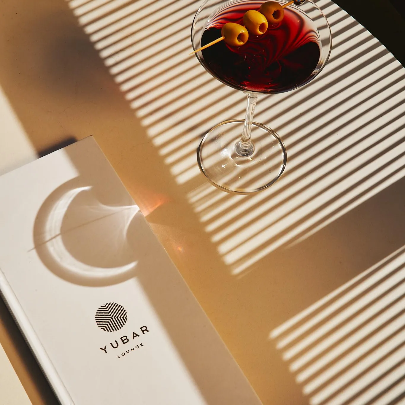 A glass of cocktail with olives and a straw, placed on a wooden table with a "YUBAR LOUNGE" menu next to it.