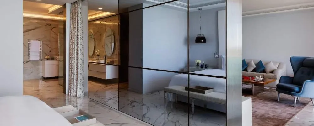 Intérieur moderne avec un miroir séparant une chambre à coucher et un salon, montrant des meubles blancs et un coin salon avec canapé et fauteuil