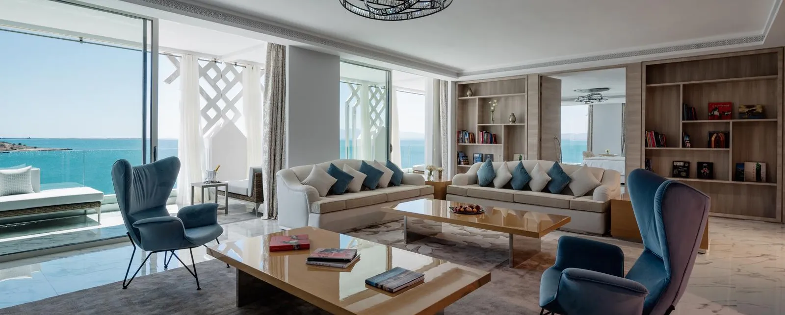 Salon moderne avec vue sur la mer, comprenant un canapé, des fauteuils, une table basse et des étagères remplies de livres