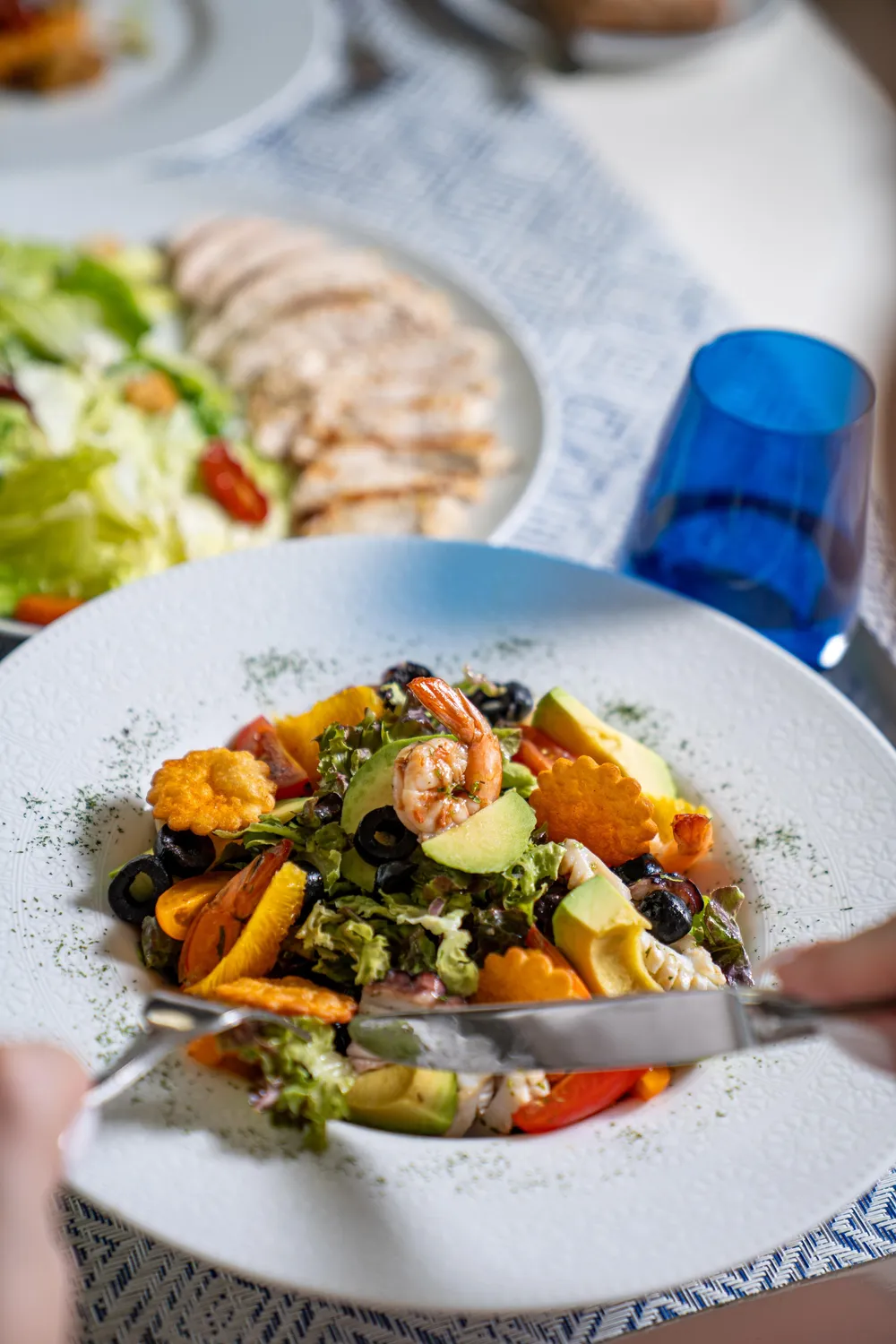 Un plato de ensalada compuesta con gambas, trozos de aguacate, aceitunas, verduras y croutones, acompañado de un tenedor