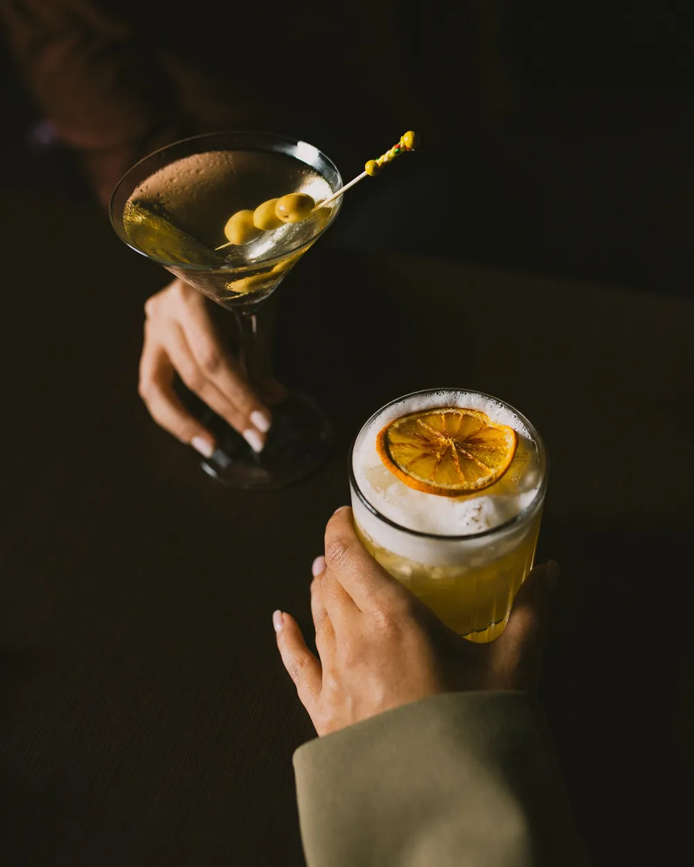 Two hands hold glasses containing drinks, one with olives and the other with a lemon slice.