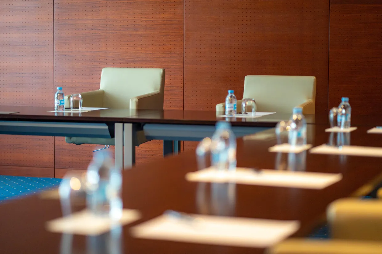 A row of conference tables with water bottles and glasses in front of empty chairs in a room with wooden walls