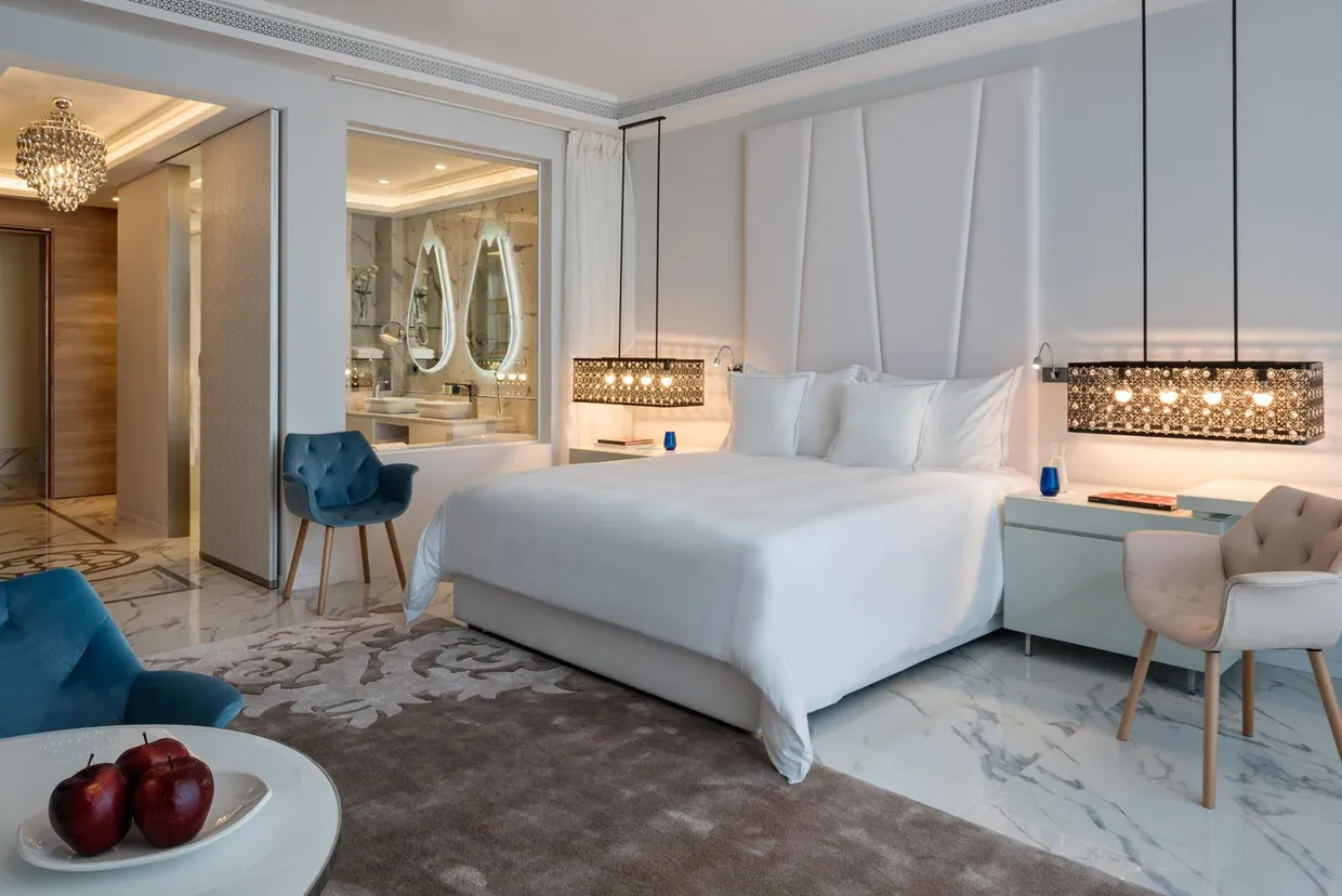 Chambre luxueuse avec lit blanc, deux fauteuils, table de chevet et vue sur une salle de bain avec miroirs ovales