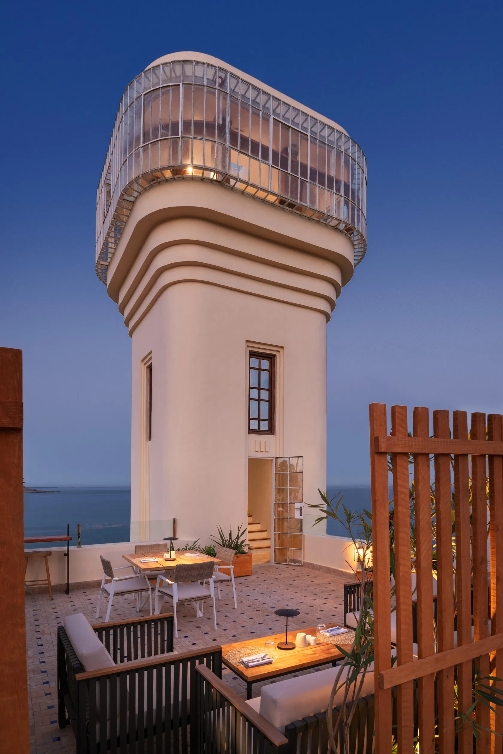 A white building with a glass-domed terrace overlooks the sea, with tables and chairs arranged on a stone terrace.