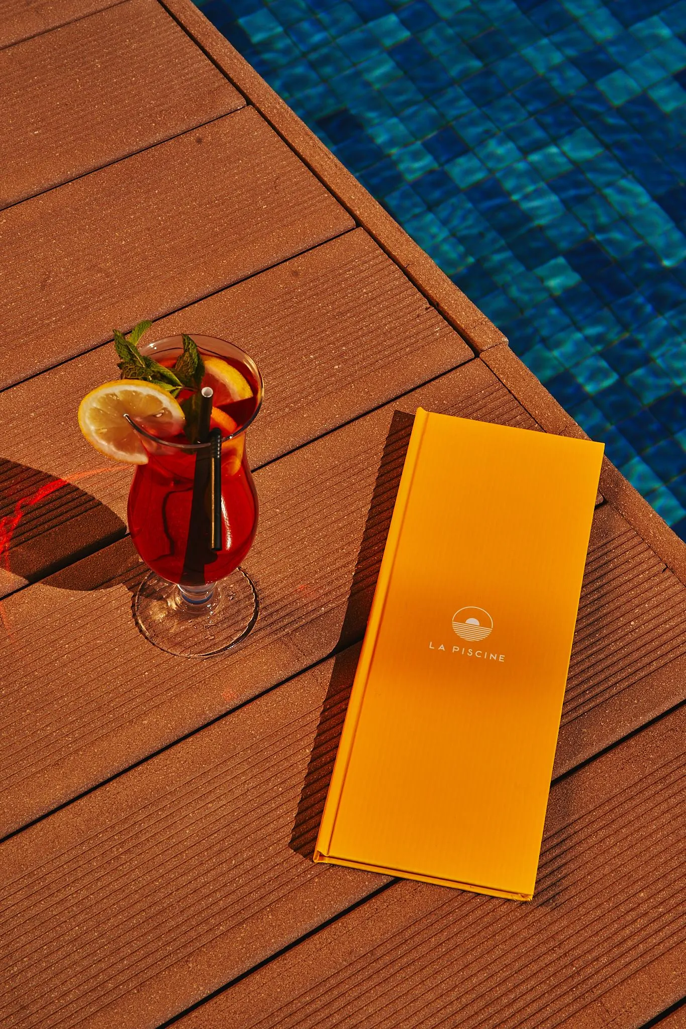 Un vaso de bebida roja con una rodaja de naranja y una pajita, un menú naranja y una piscina azul de fondo