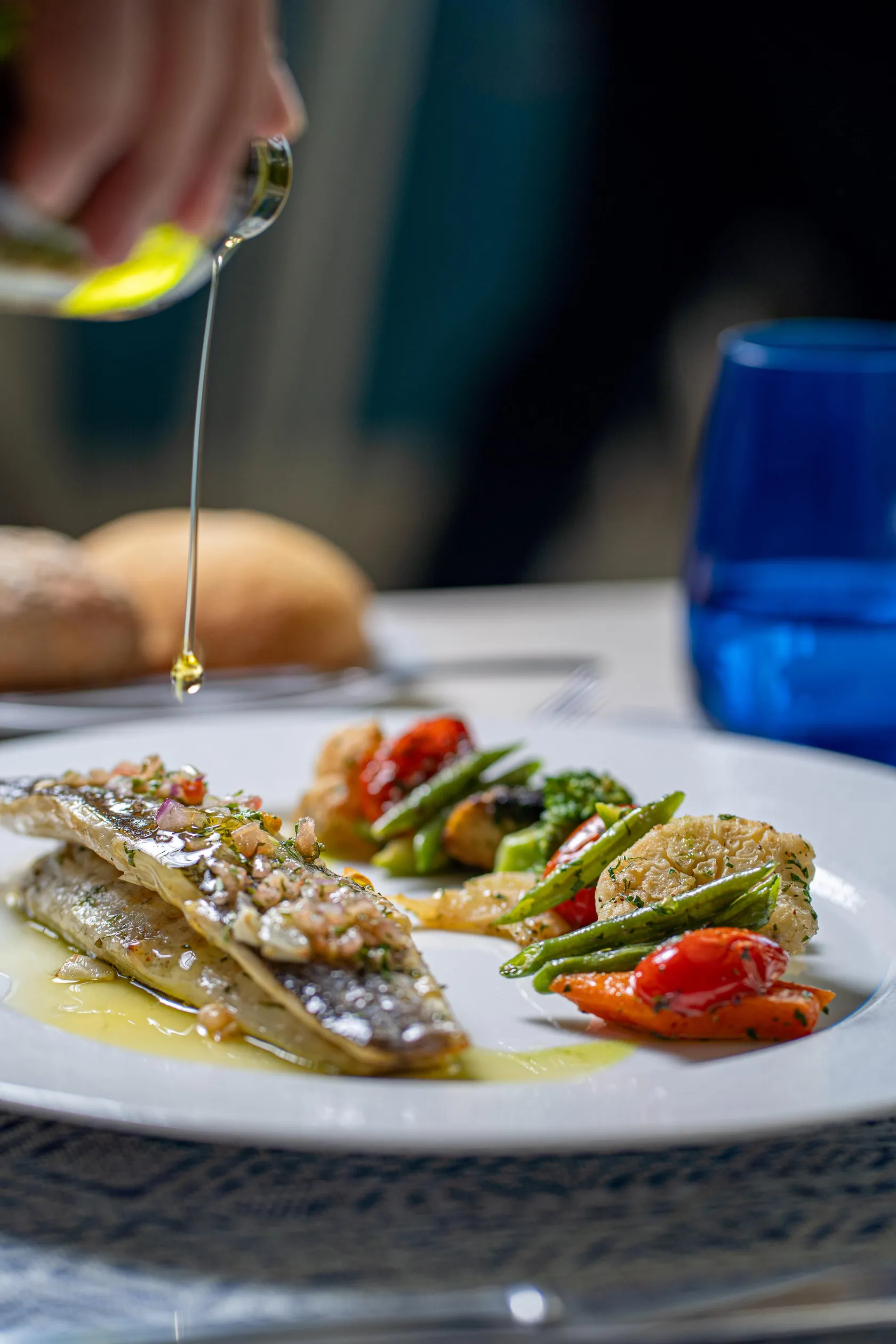 Una mano vierte aceite sobre un plato compuesto de pescado y verduras a la parrilla, acompañado de pan.