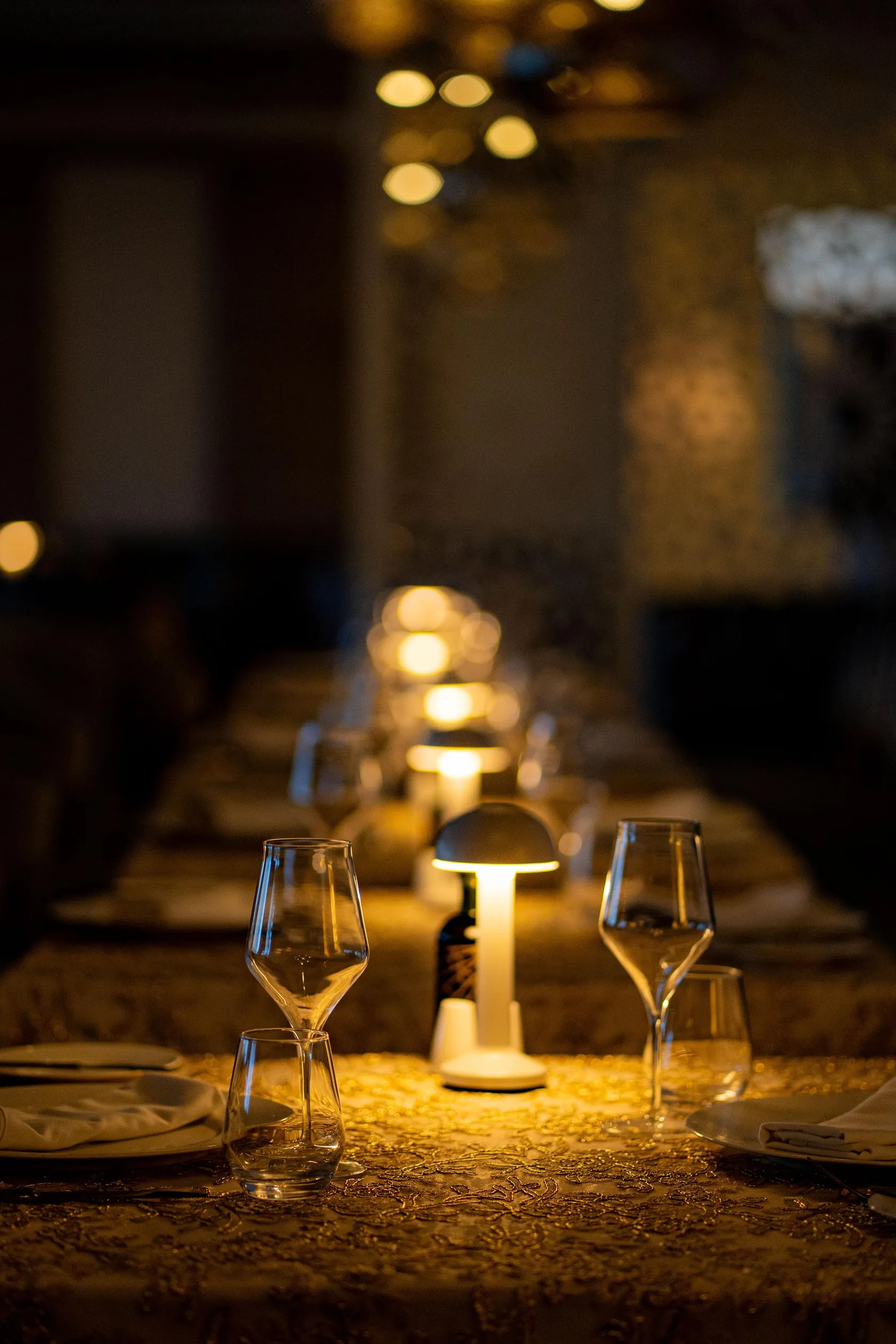 Mesa preparada con copas de vino, mantel y velas, iluminada por lámparas en el fondo