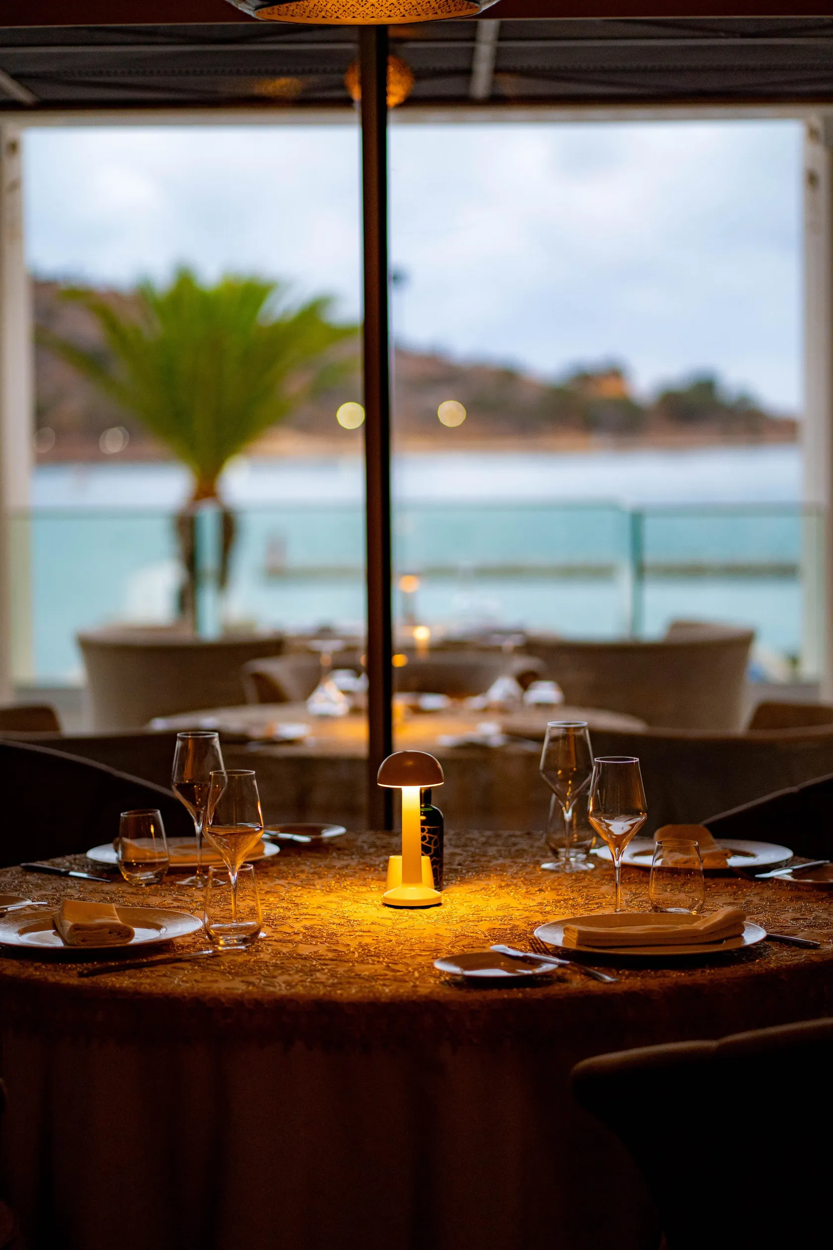 Table set with wine glasses and lit candle, seen through a window overlooking a terrace and an outdoor landscape with palm trees and sea.