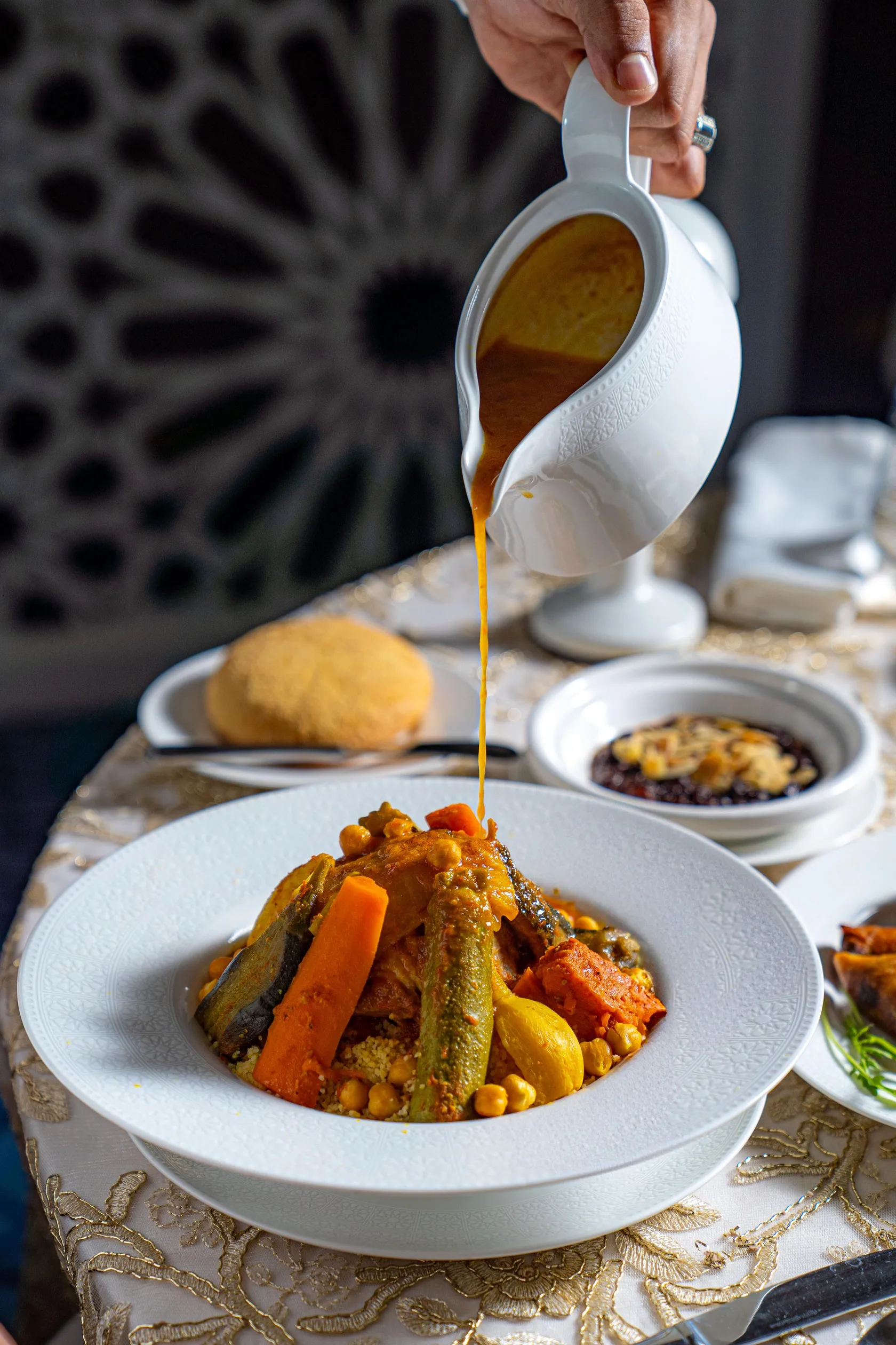 A hand pours sauce from a ladle onto a dish containing vegetables and chickpeas.