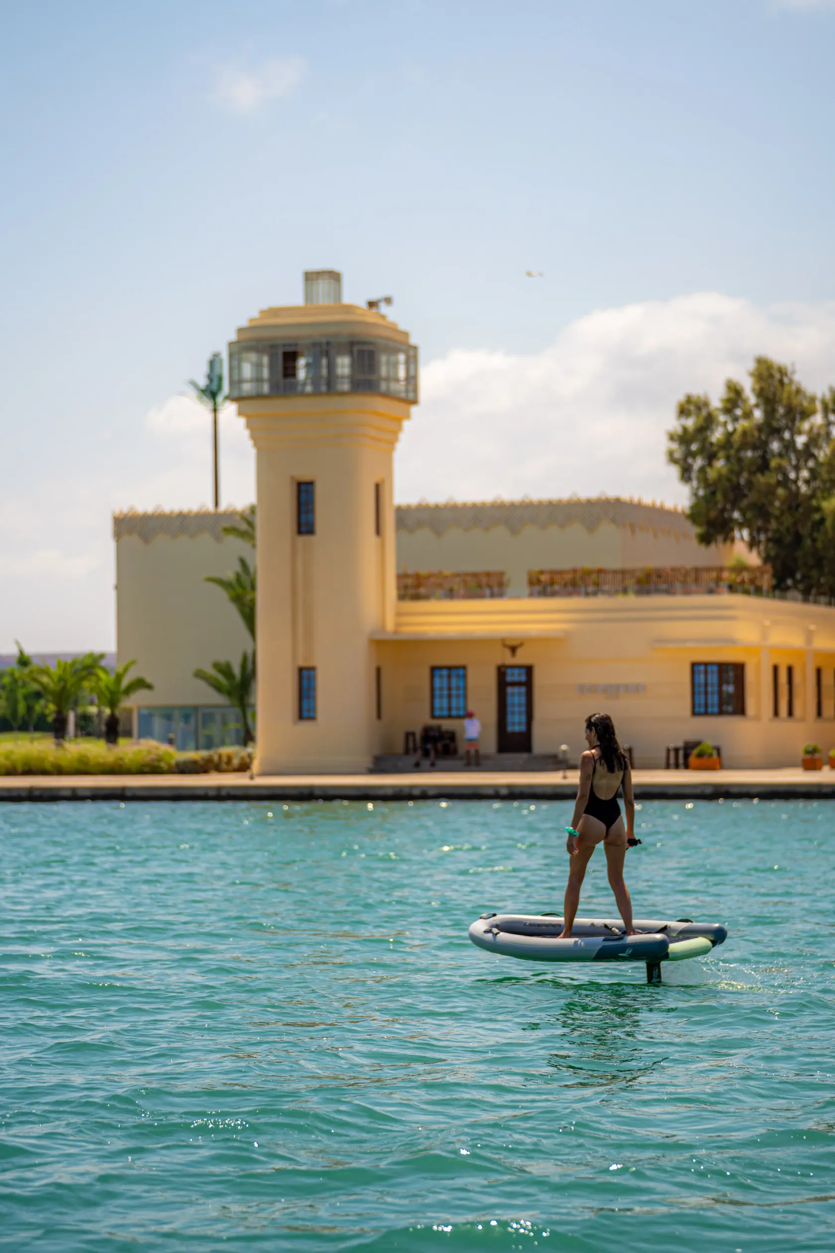 Una persona con traje de baño hace paddle en una extensión de agua frente a un edificio amarillo con una torre.