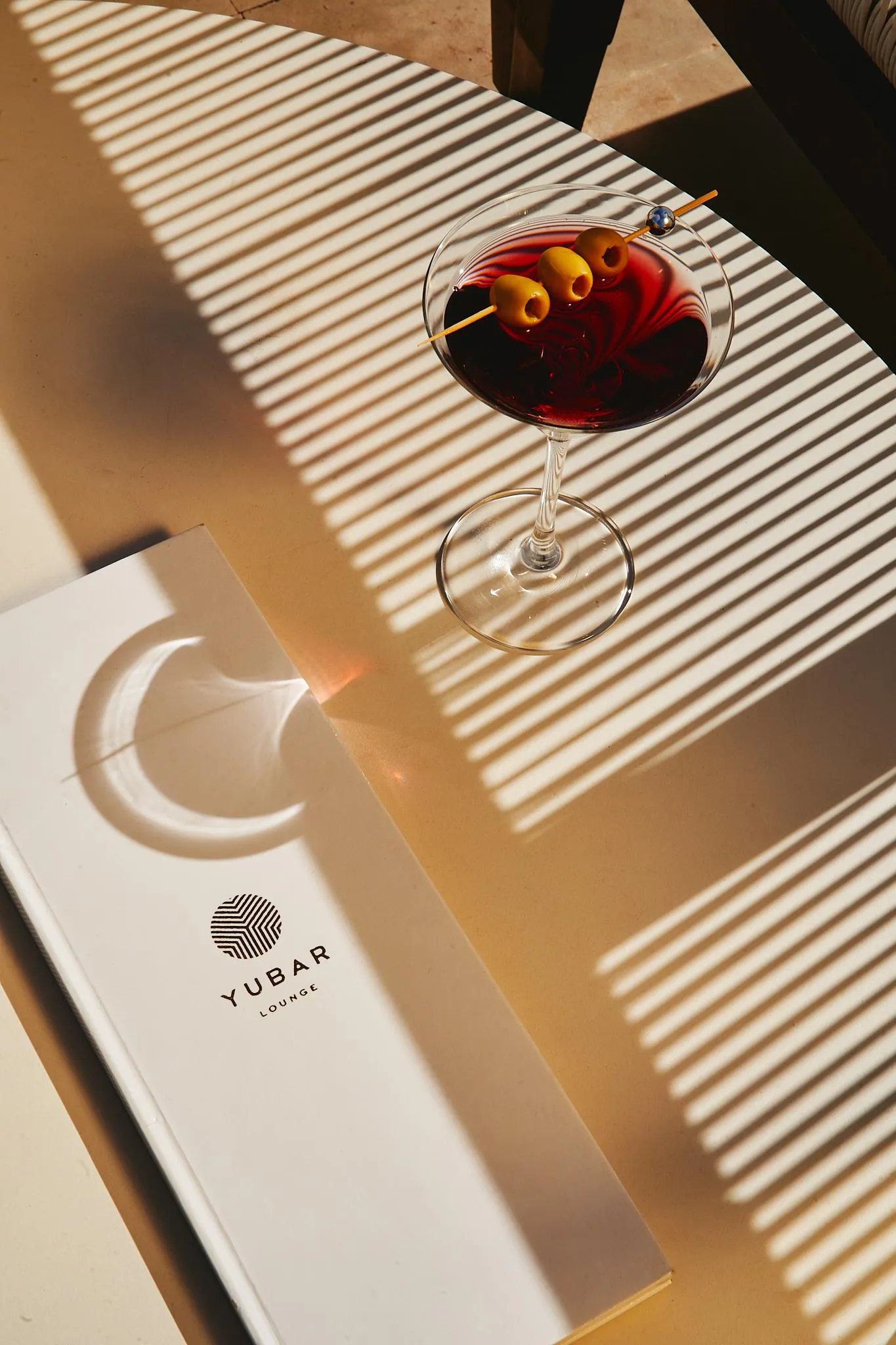 Un vaso de cóctel con aceitunas y una pajita, colocado sobre una mesa de madera con un menú "YUBAR LOUNGE" al lado