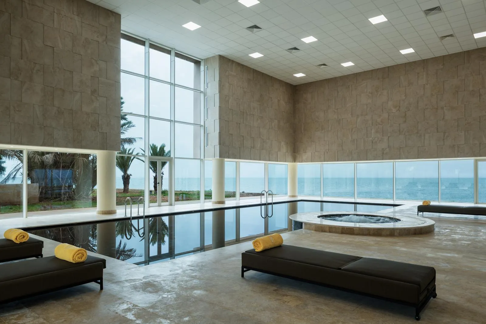 Piscine intérieure avec vue sur la mer, entourée de canapés et de serviettes jaunes