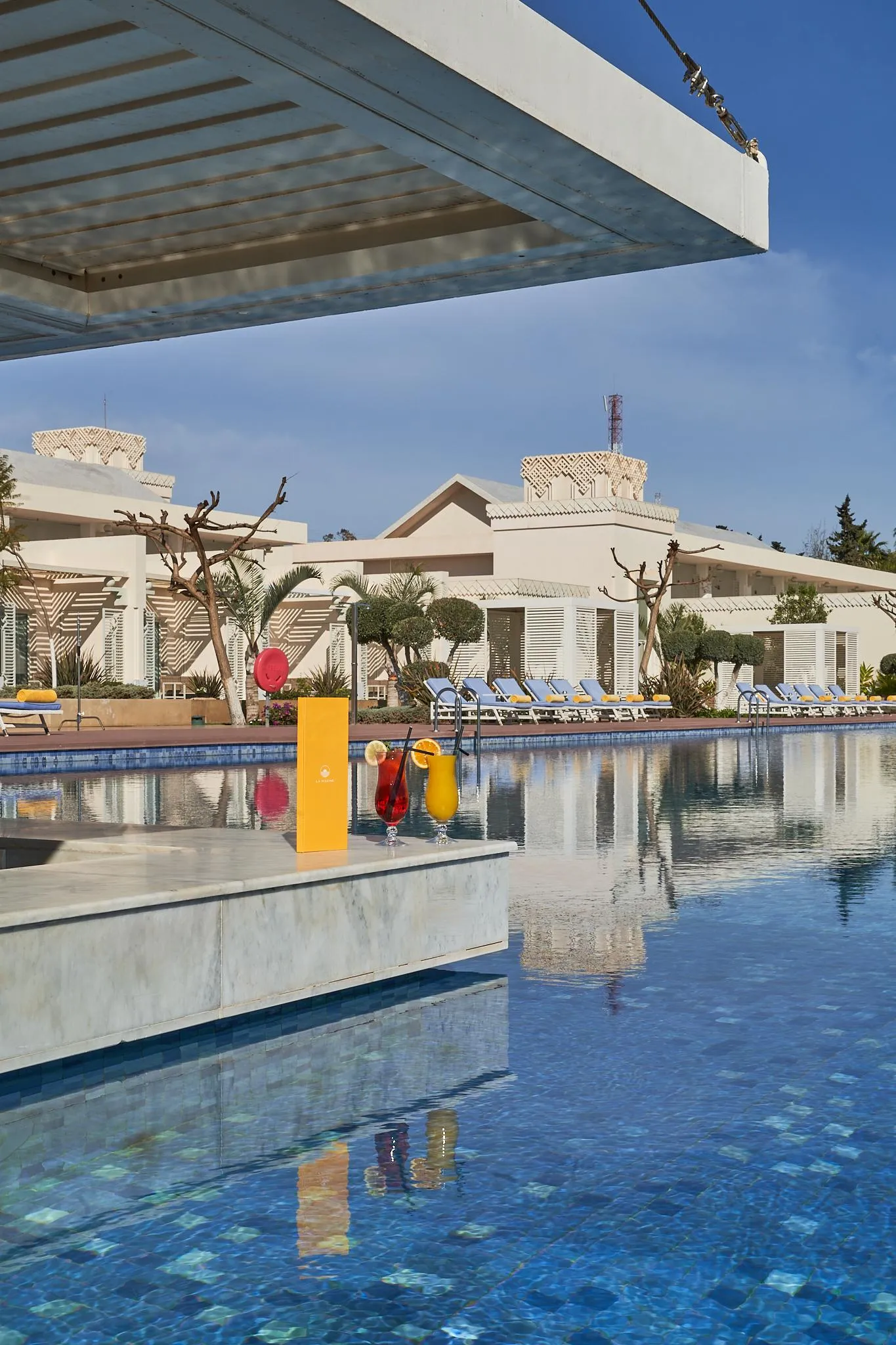 Pool with two glasses of colorful drinks on the edge, surrounded by sun loungers and white buildings with flat roofs