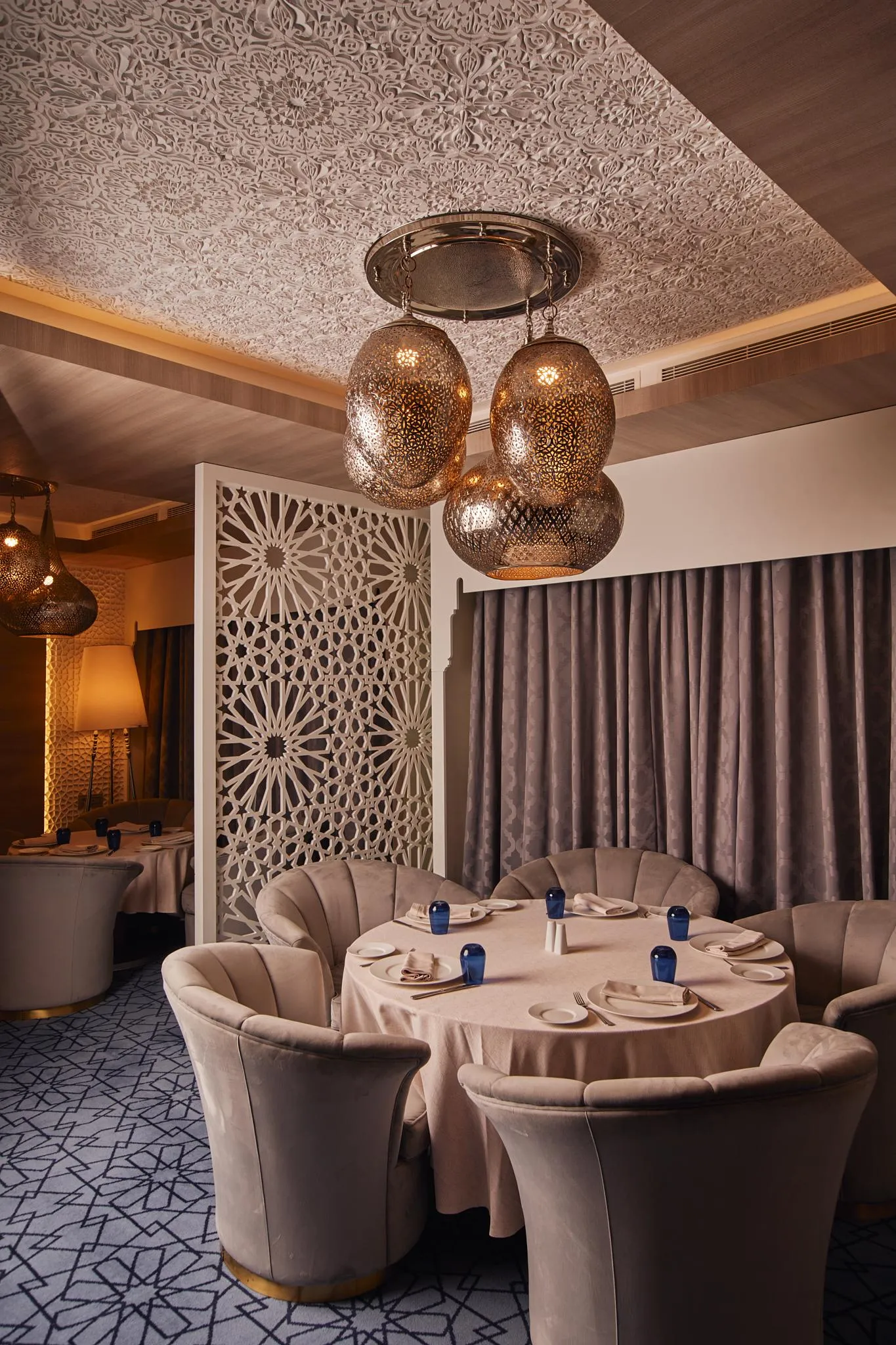 Un restaurante elegante con mesas redondas, sillas acolchadas de terciopelo beige, lámparas de metal y un techo decorado con motivos geométricos