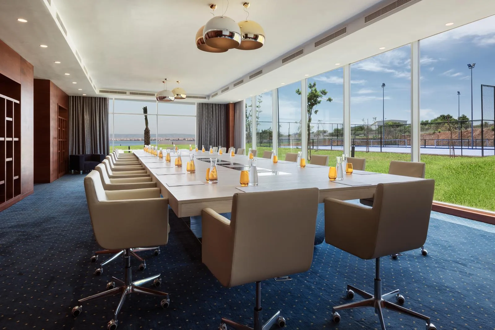 Salle de réunion avec une longue table entourée de chaises, des verres et des bougeoirs sur la table, de grandes fenêtres offrant une vue sur un terrain extérieur