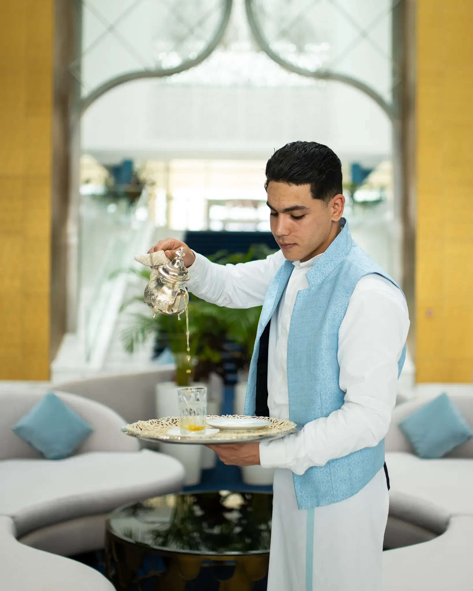 Un serveur en tenue blanche et bleu verse de l'eau dans un verre posé sur un plateau