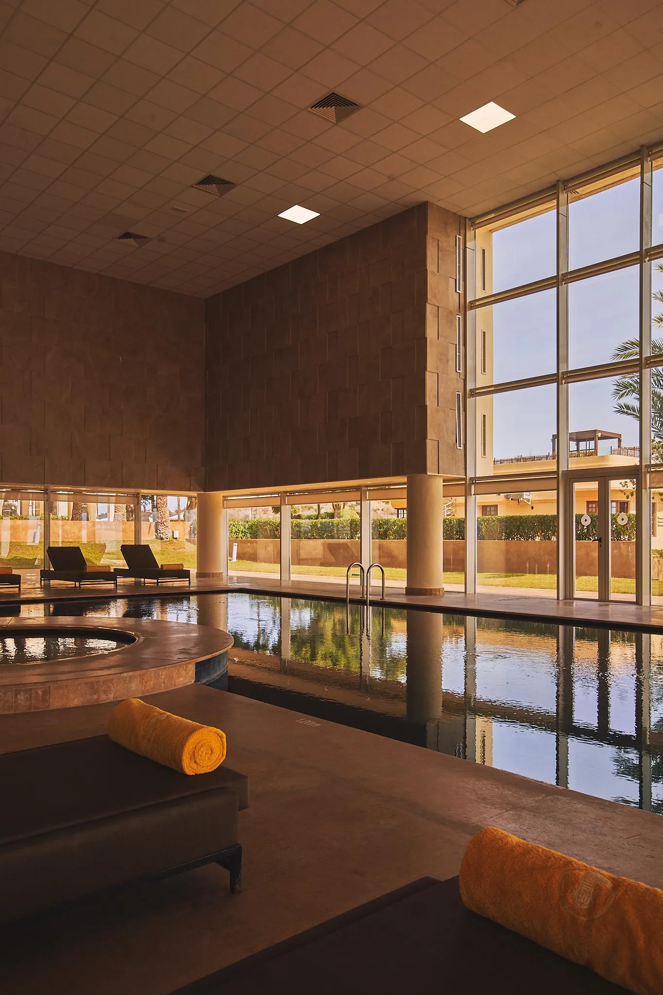 Piscina interior con tumbonas y toallas, rodeada de grandes ventanas y muros de piedra