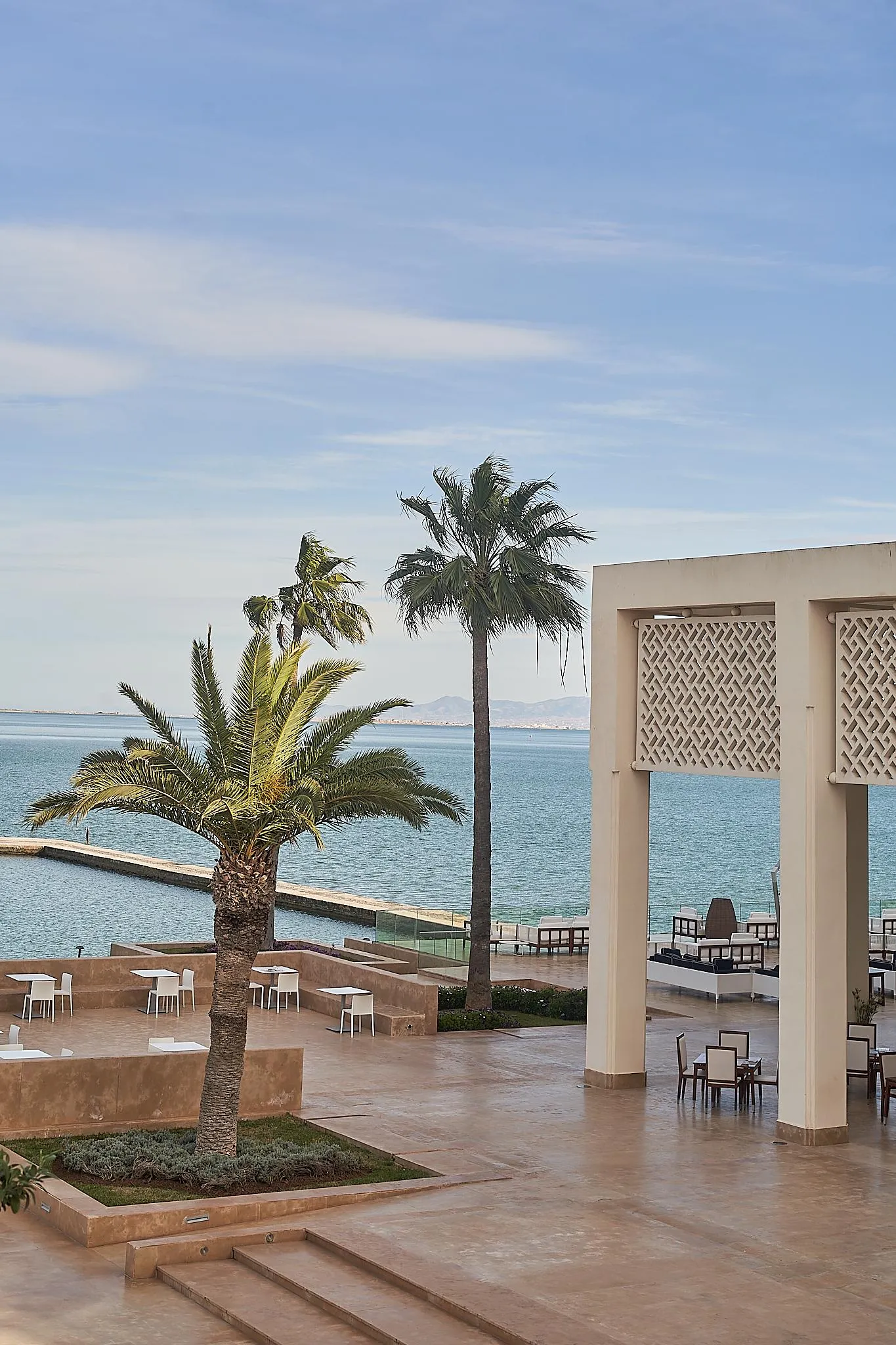 Terraza con mesas y sillas cerca del mar, bordeada de palmeras y una estructura arquitectónica con motivos geométricos