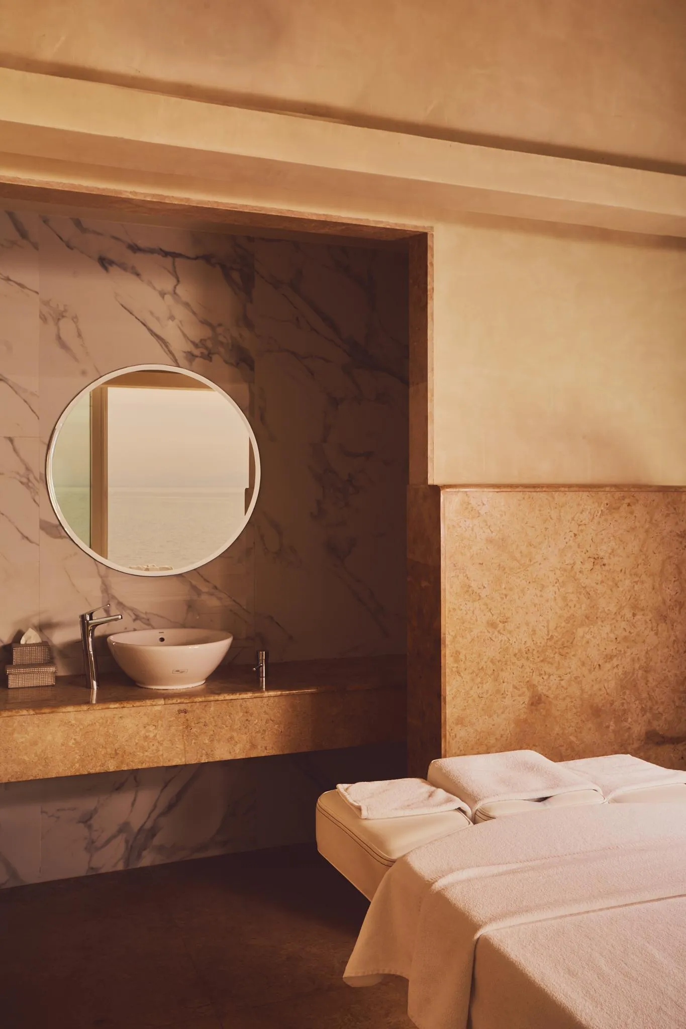 Un lavabo encastré dans un plan de travail en pierre avec un miroir rond au-dessus, à côté d'une table de massage recouverte de serviettes blanches