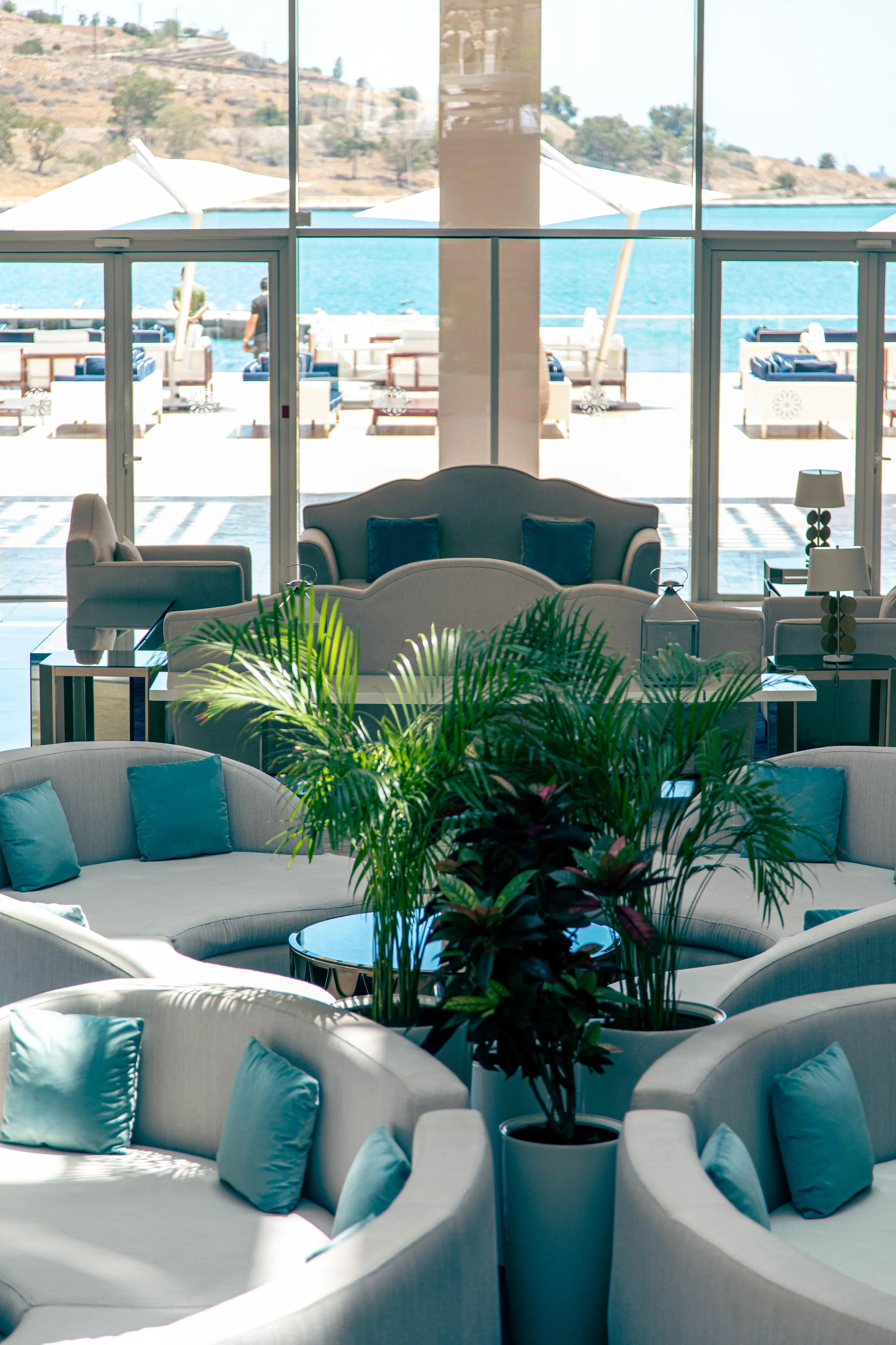 Un salón con sofás grises y cojines azules, una planta verde en el centro, y una vista a una piscina y el mar a través de grandes ventanales.