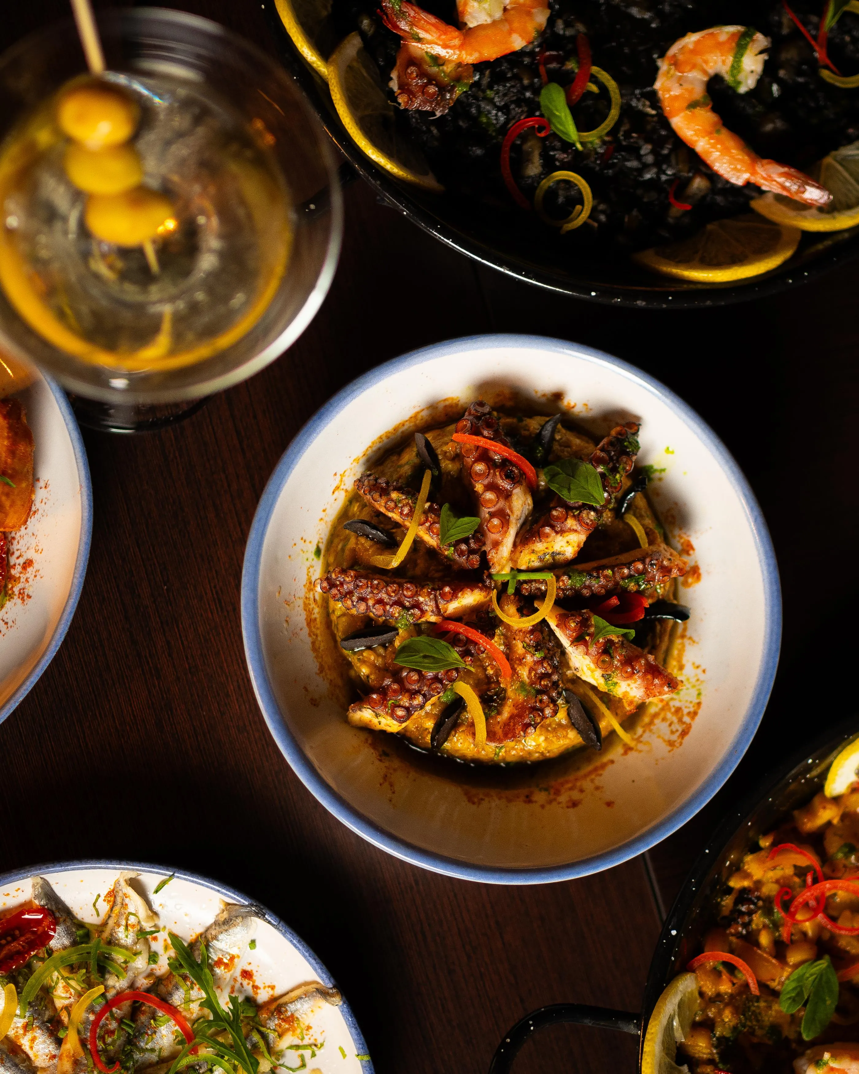 A grilled squid dish with vegetables and herbs, served with other seafood plates and a drink in a glass.