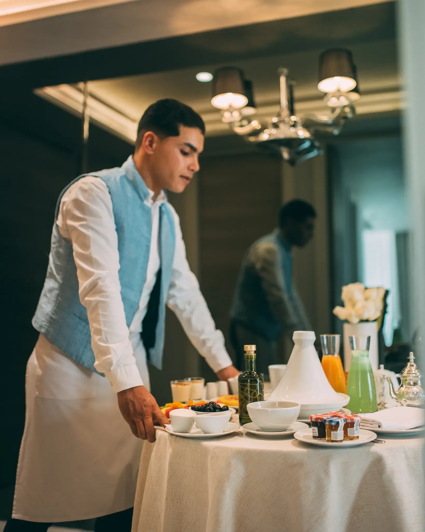 Un serveur en veste bleue et tablier blanc prépare une table avec des bols de fruits, des boissons et des petits pots de confiture