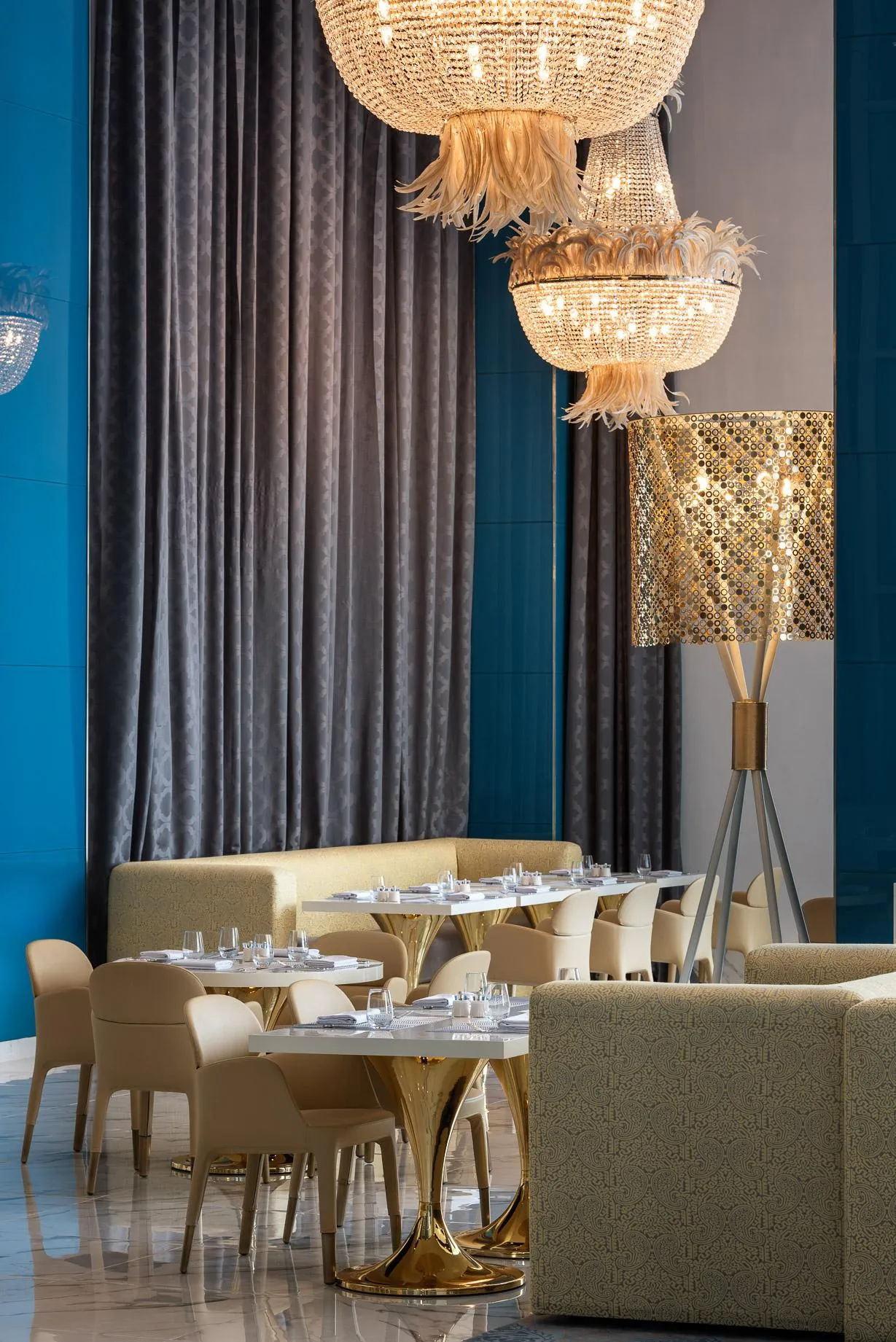 Sala de restaurante elegante con mesas redondas, sillas y sofás beige, lámparas de cristal y cortinas grises