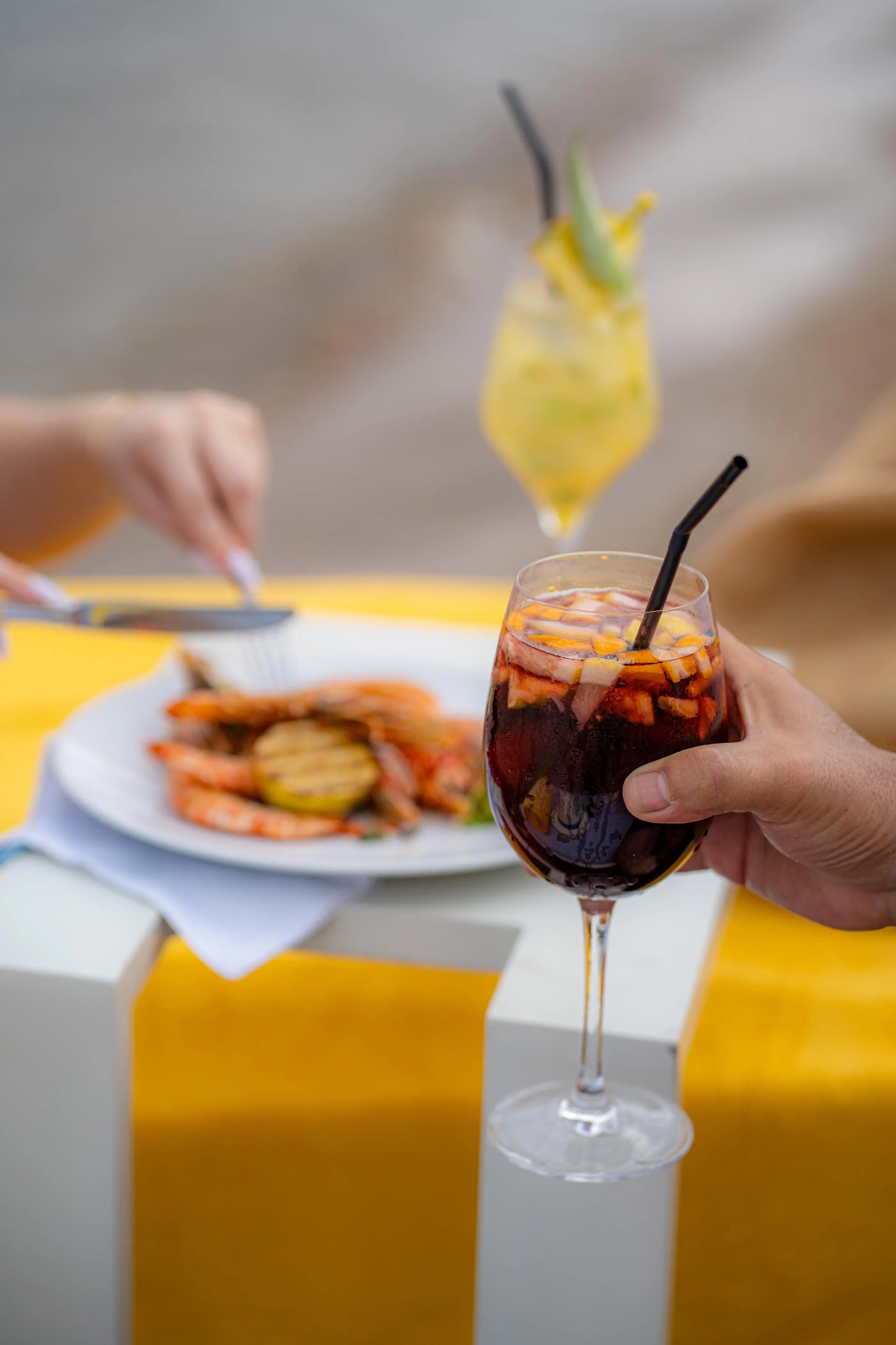 Una persona sostiene un vaso de bebida roja con hielo y una pajilla, frente a un plato de comida y una mesa amarilla.