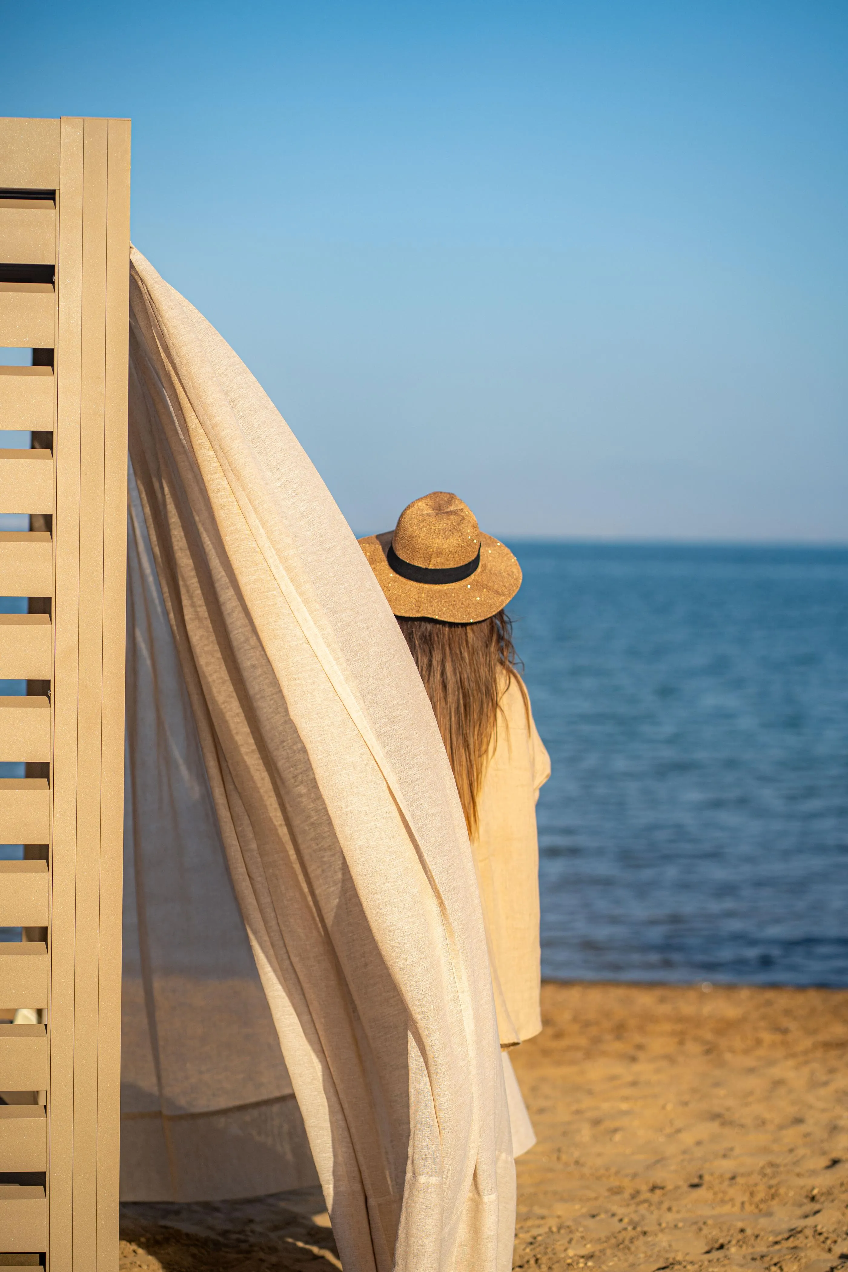 Una persona con sombrero mira el mar desde una playa, una sábana blanca colgada de una estructura de madera a su lado.